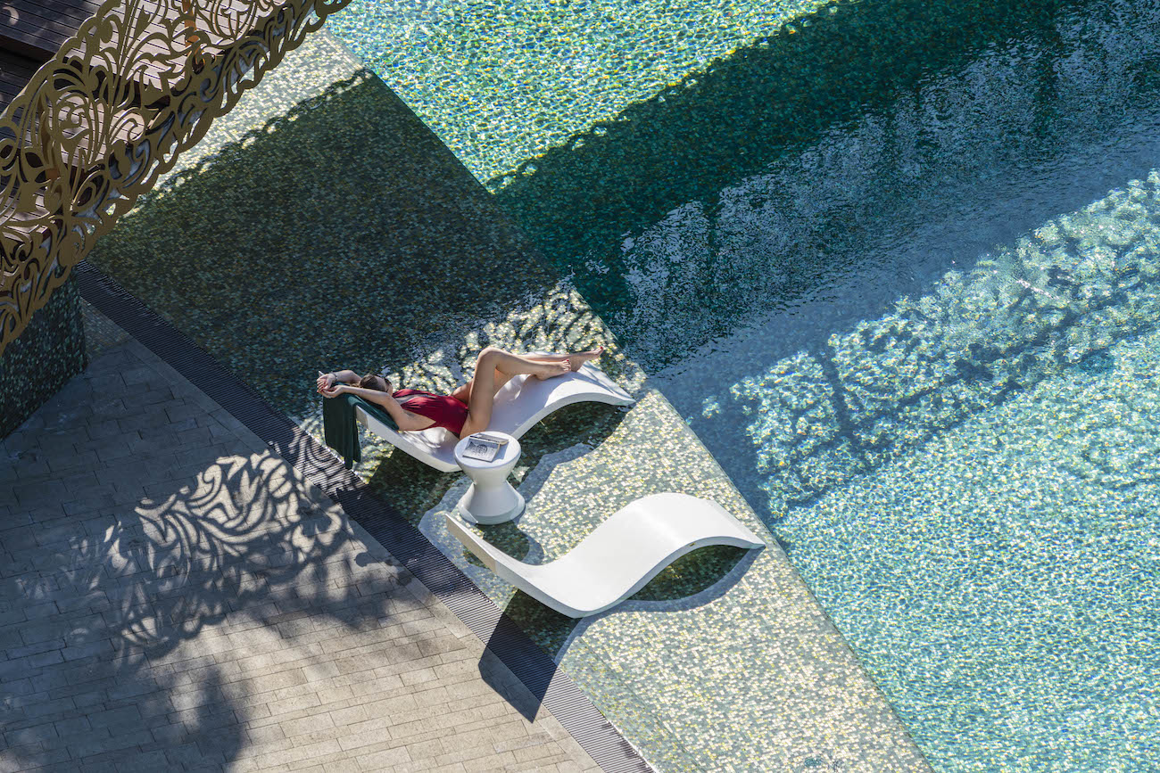A person in a red swimsuit lounges on a white chair beside a tiled pool, with a small table nearby and decorative shadows on the ground.