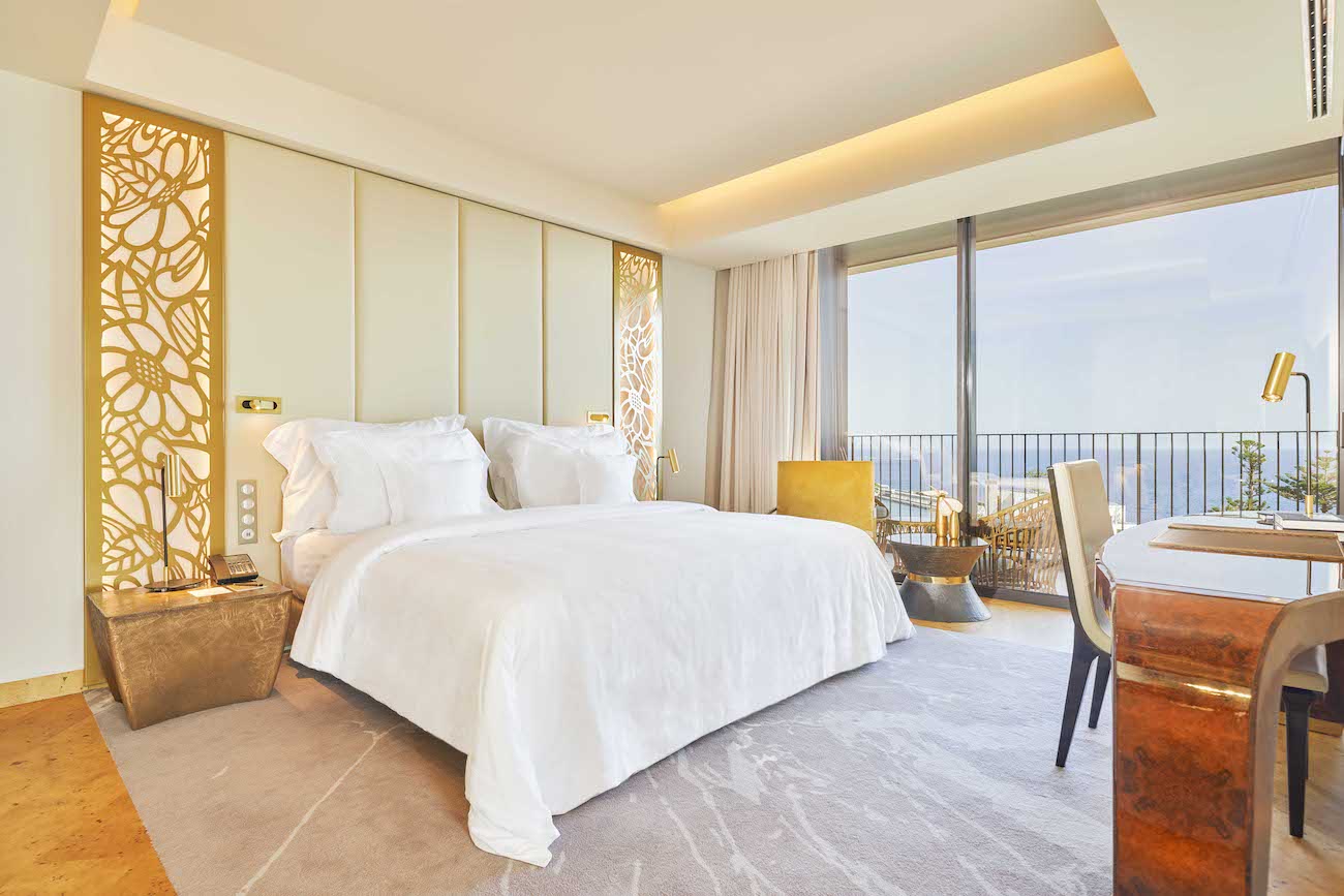 Modern hotel room with a large bed, decorative gold panels, desk, and floor-to-ceiling windows opening to a balcony with outdoor seating and a scenic view.