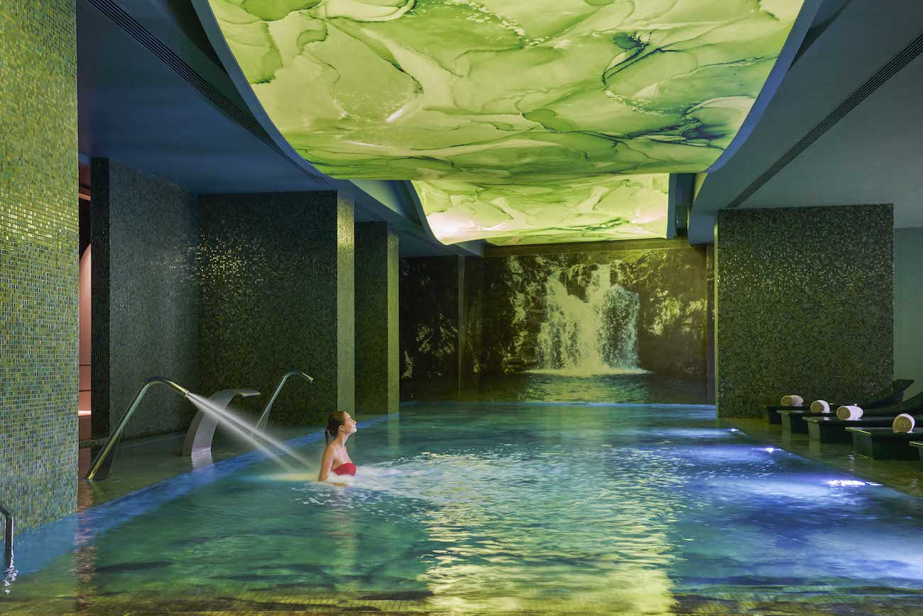 A woman relaxes in an indoor pool with mosaic tile walls, water features, lounge chairs, and a green, abstract ceiling design.