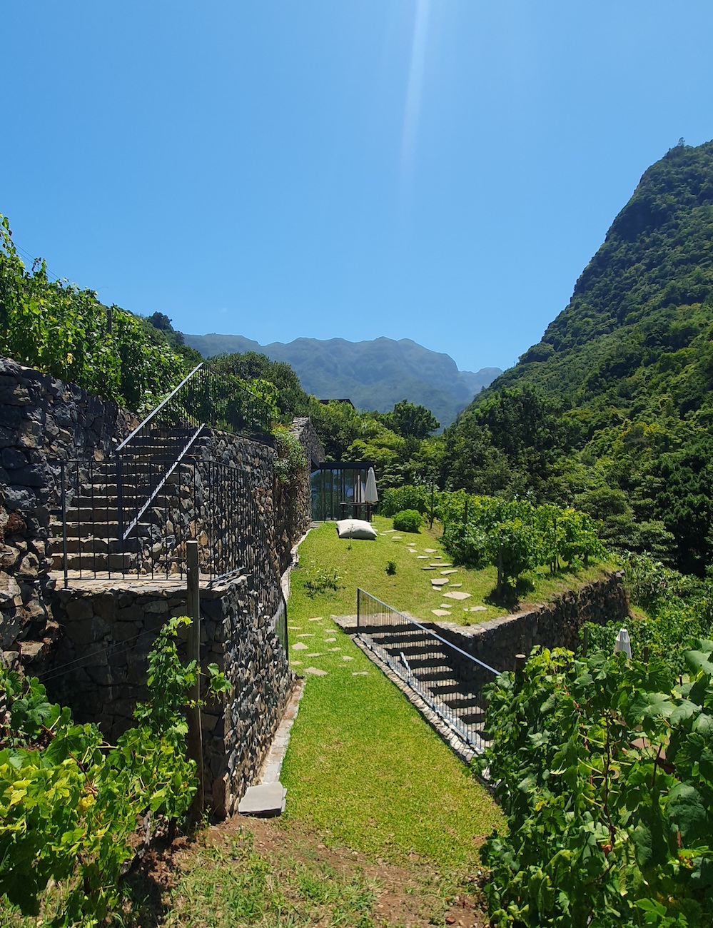 Stone staircases and pathways border a grassy area with garden plants, set between lush green mountains under a clear blue sky.