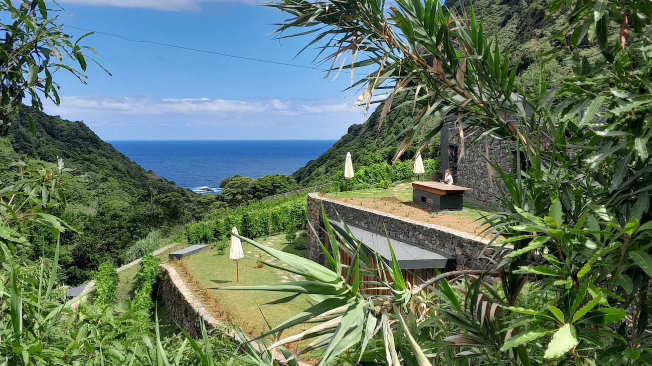 Modern stone building with grass roof and patio umbrellas overlooks lush green valley and blue ocean under a partly cloudy sky, surrounded by dense tropical vegetation.