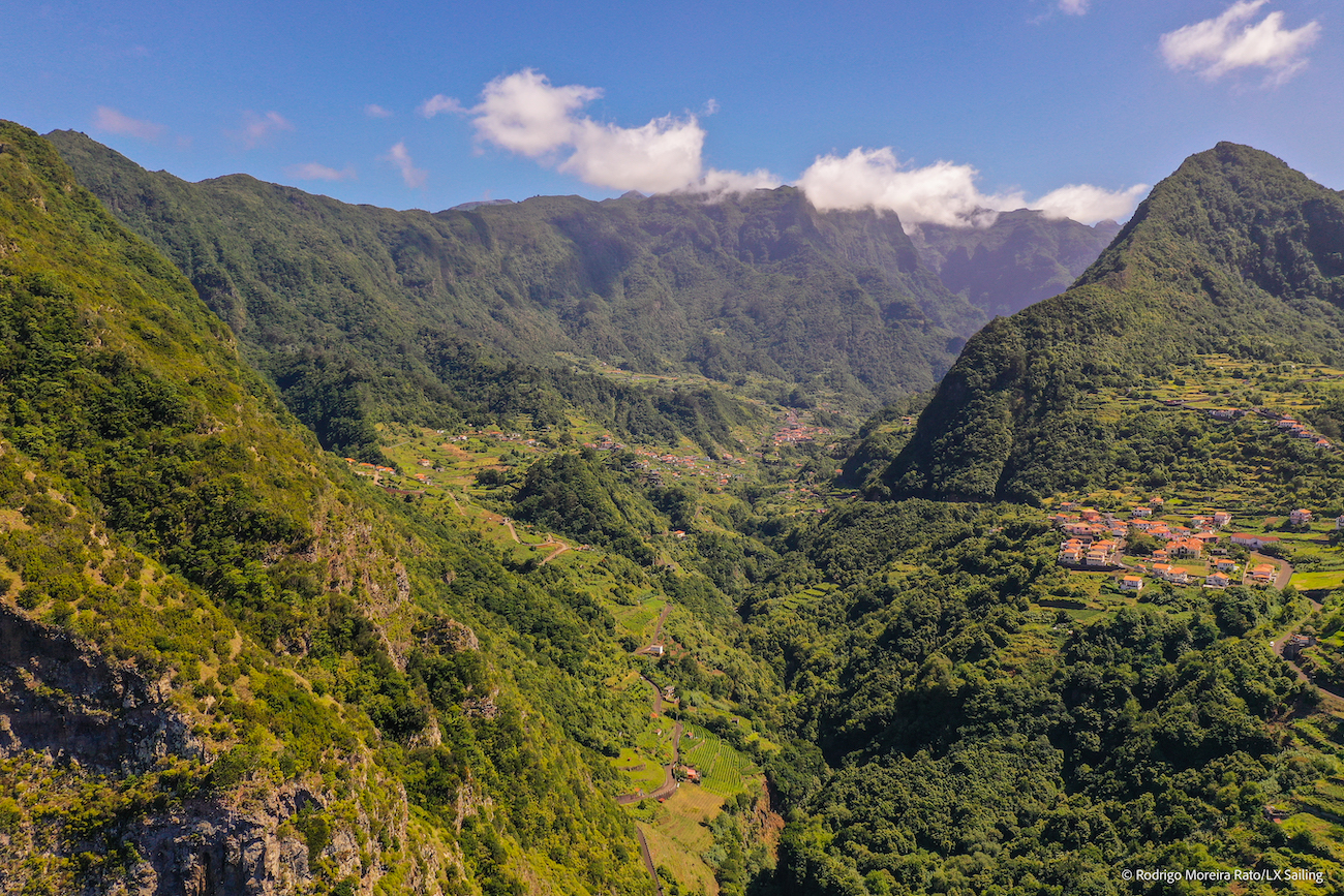 Aerial view of a lush green valley surrounded by steep mountains, with small villages and terraced fields under a partly cloudy sky.