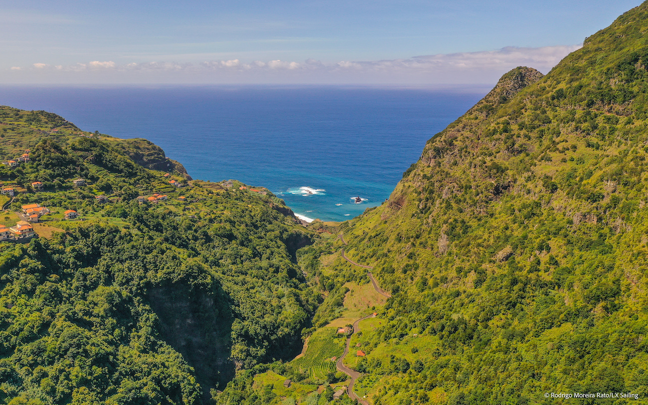 Aerial view of a green valley with scattered houses leading to the blue ocean, bordered by steep, forested hills under a clear sky.