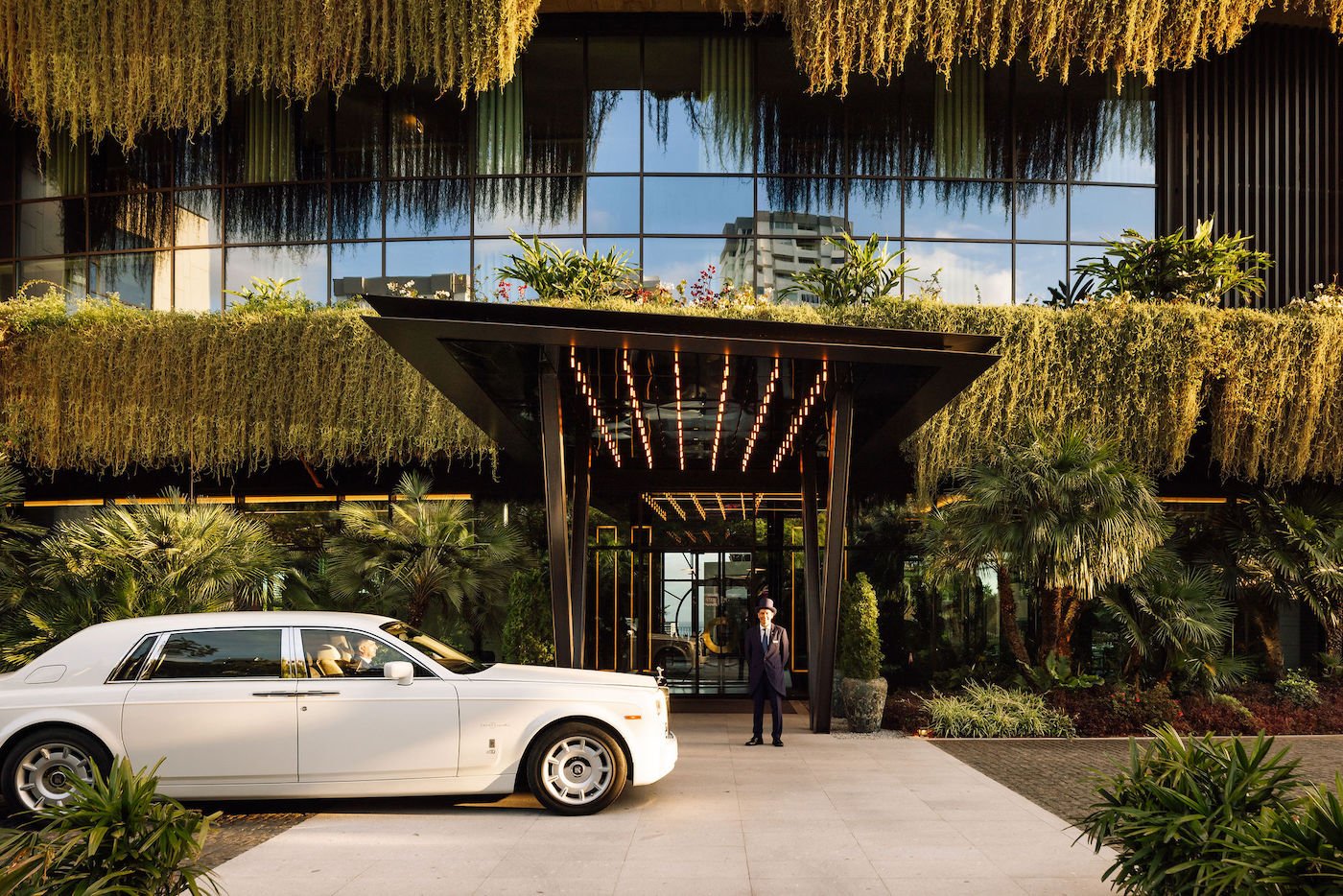 A white luxury car is parked in front of a modern building with lush hanging greenery and a doorman standing at the entrance.