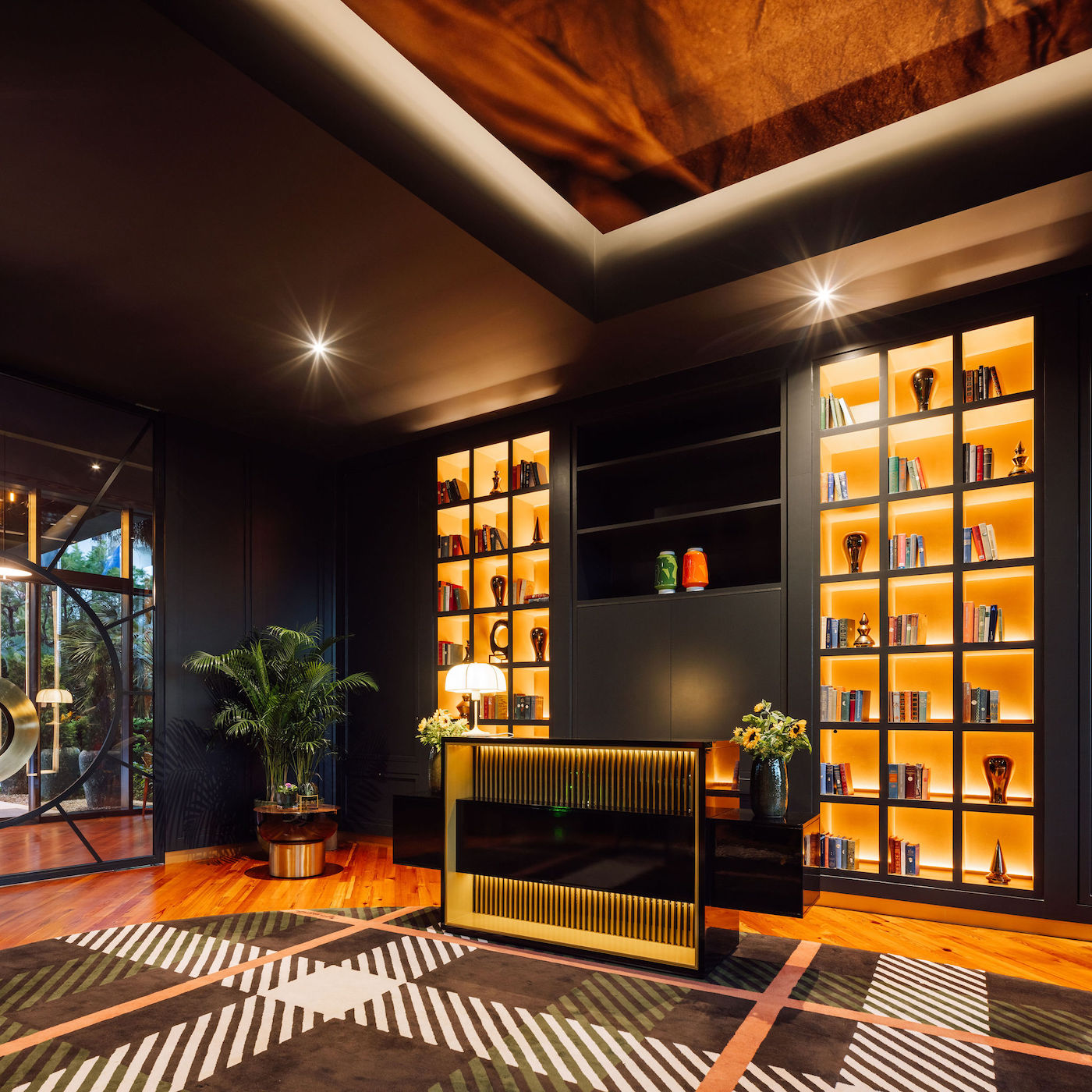 Modern home library with illuminated bookshelves, a black and gold desk, potted plants, and geometric patterned rug under warm lighting.
