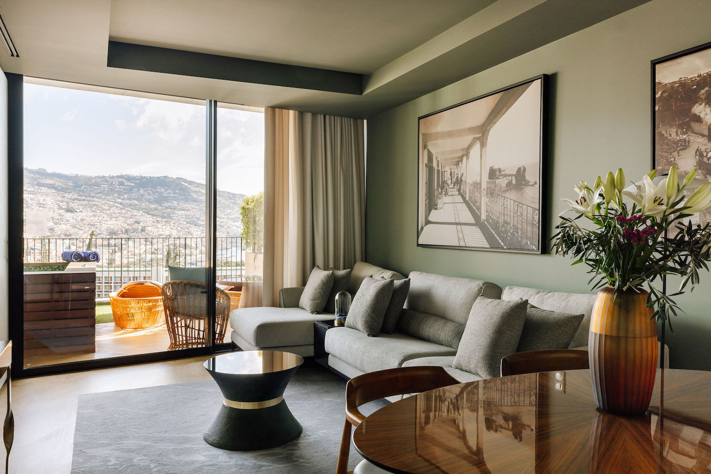 Modern living room with a sectional sofa, coffee table, large artwork, and a vase of flowers, opening to a balcony with outdoor seating and a scenic view through glass doors.