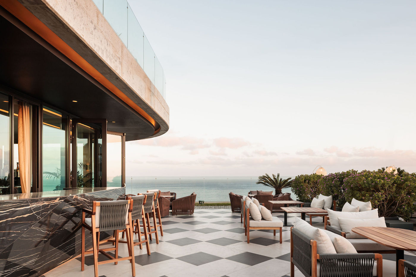 Outdoor restaurant patio with modern furnishings, a marble bar, and a view overlooking the ocean, under a clear sky.