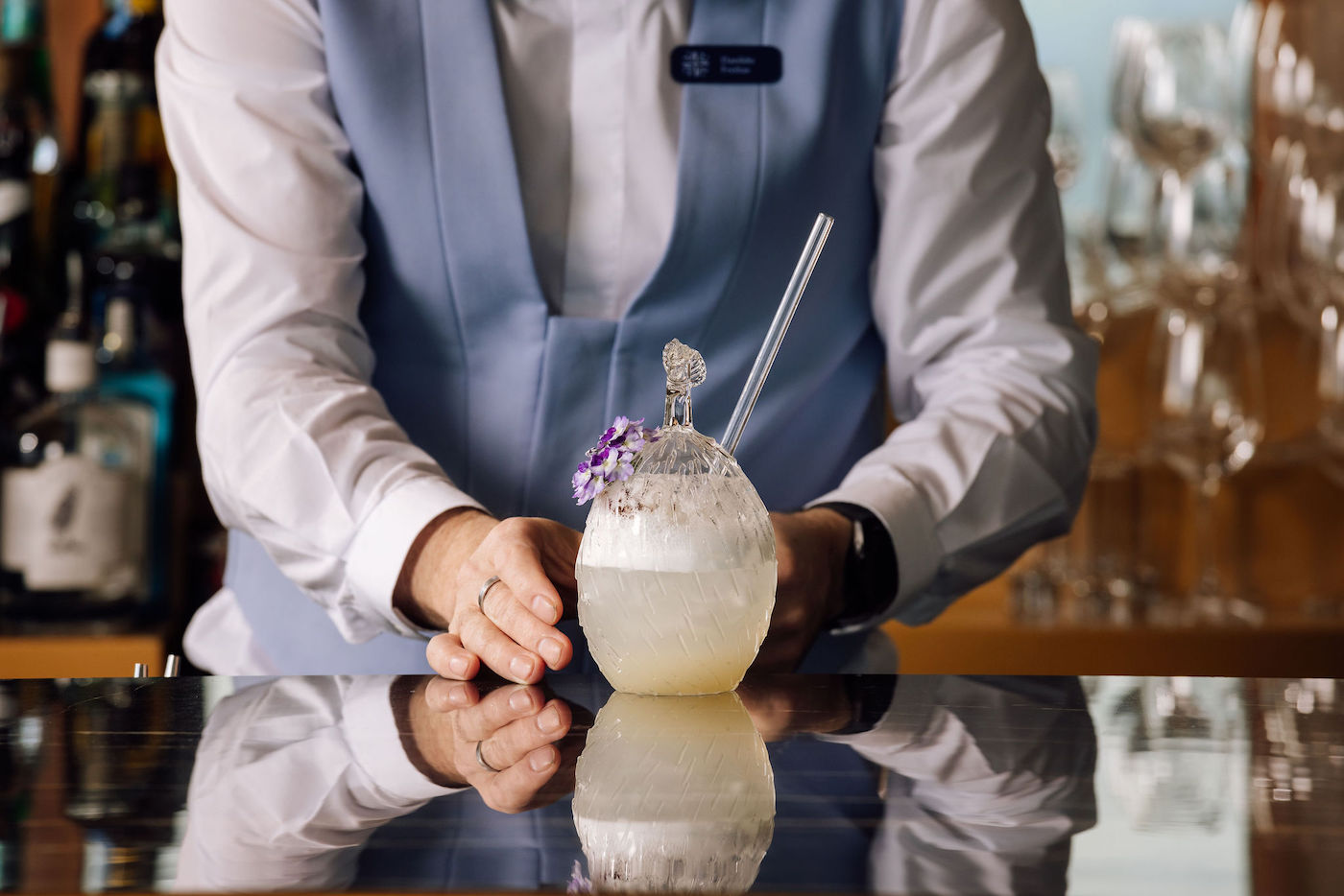 A bartender in a light blue vest presents a round cocktail garnished with a flower and served with a straw on a reflective bar counter.