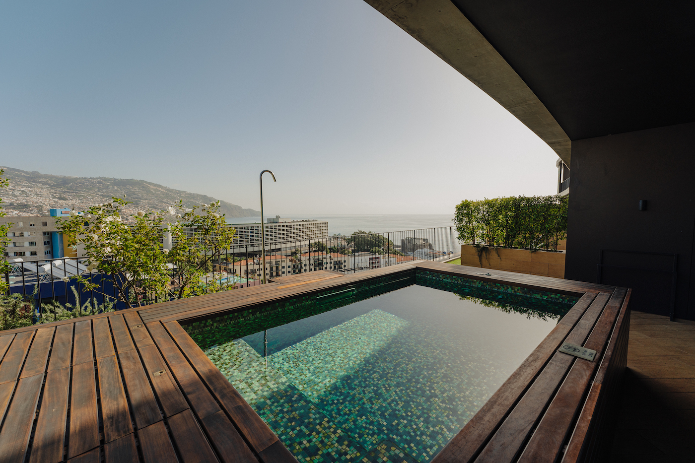 A small rectangular pool with wooden decking overlooks a cityscape and the ocean under a clear sky.