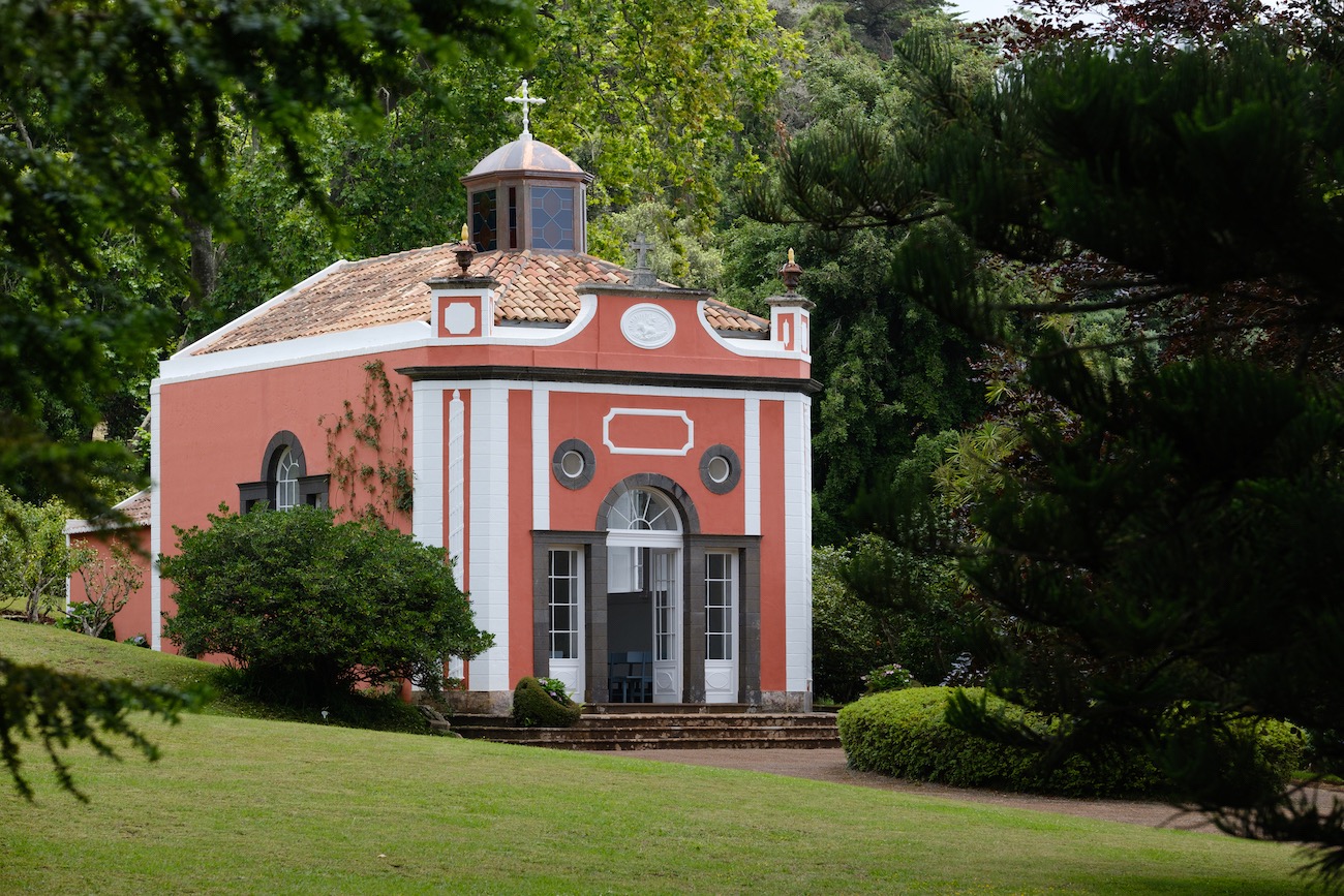 A small, red and white chapel with arched windows and a cross on top stands surrounded by green trees and manicured bushes.