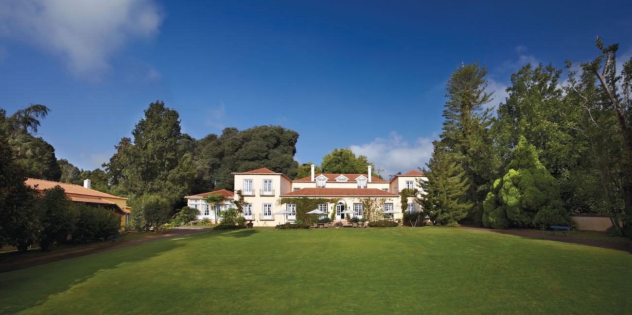 Large white house with red roof sits behind a wide, well-kept lawn, surrounded by tall trees and greenery under a blue sky.