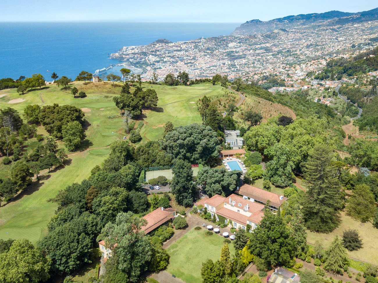 Aerial view of a hillside estate with red-roofed buildings, gardens, and a pool, overlooking a city by the sea with mountains in the background.