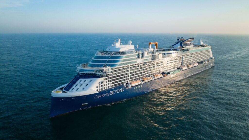 A large blue and white cruise ship sails on calm ocean waters under a clear sky.