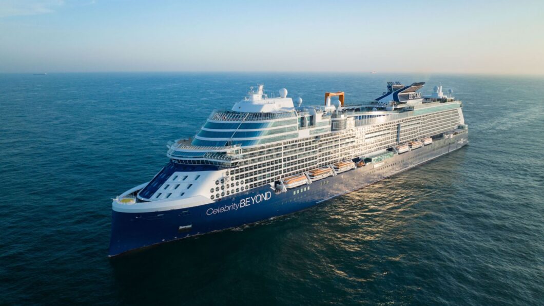 A large blue and white cruise ship sails on calm ocean waters under a clear sky.
