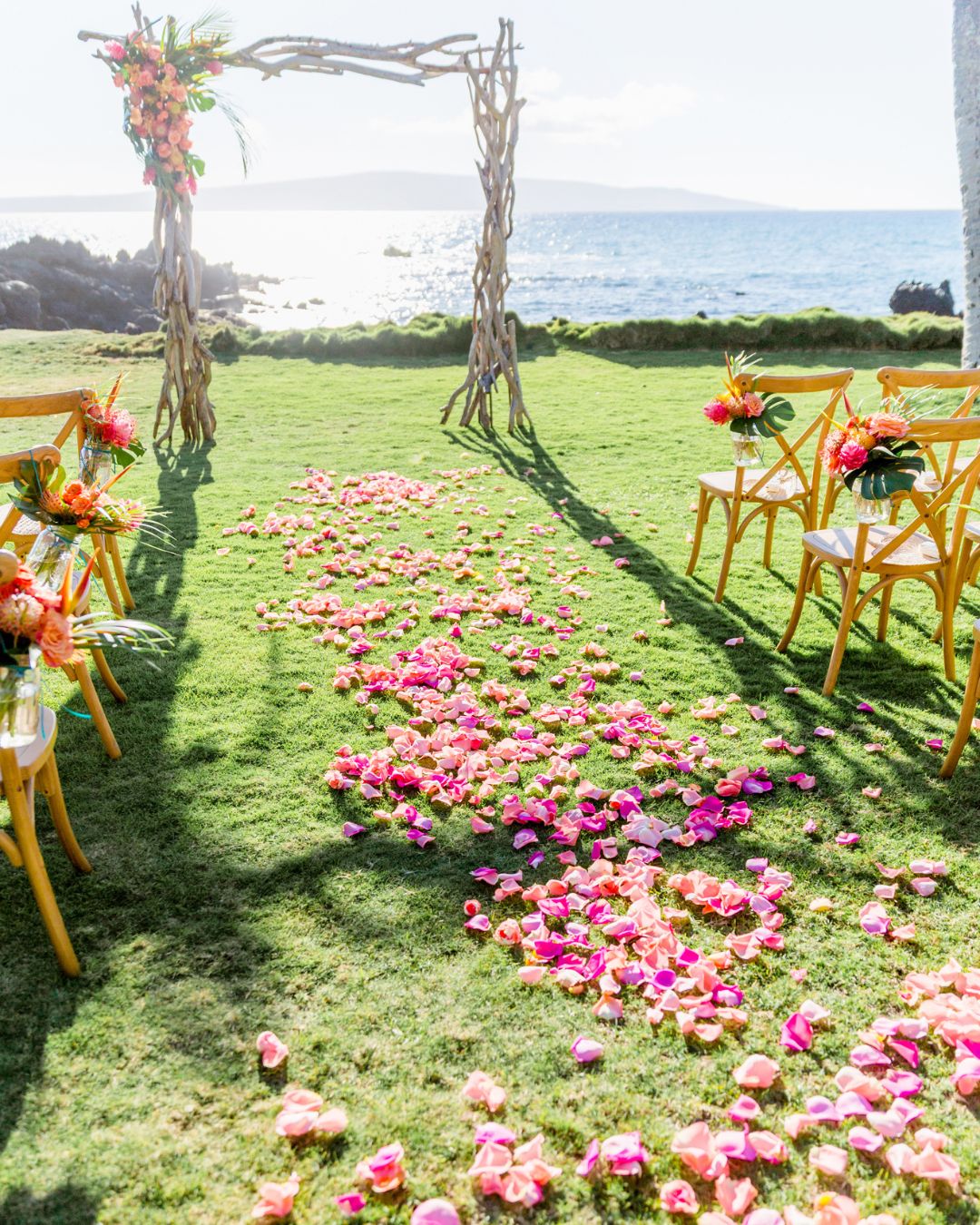 Outdoor wedding setup with wooden arch, yellow chairs, and a path of pink flower petals on green grass by the ocean.
