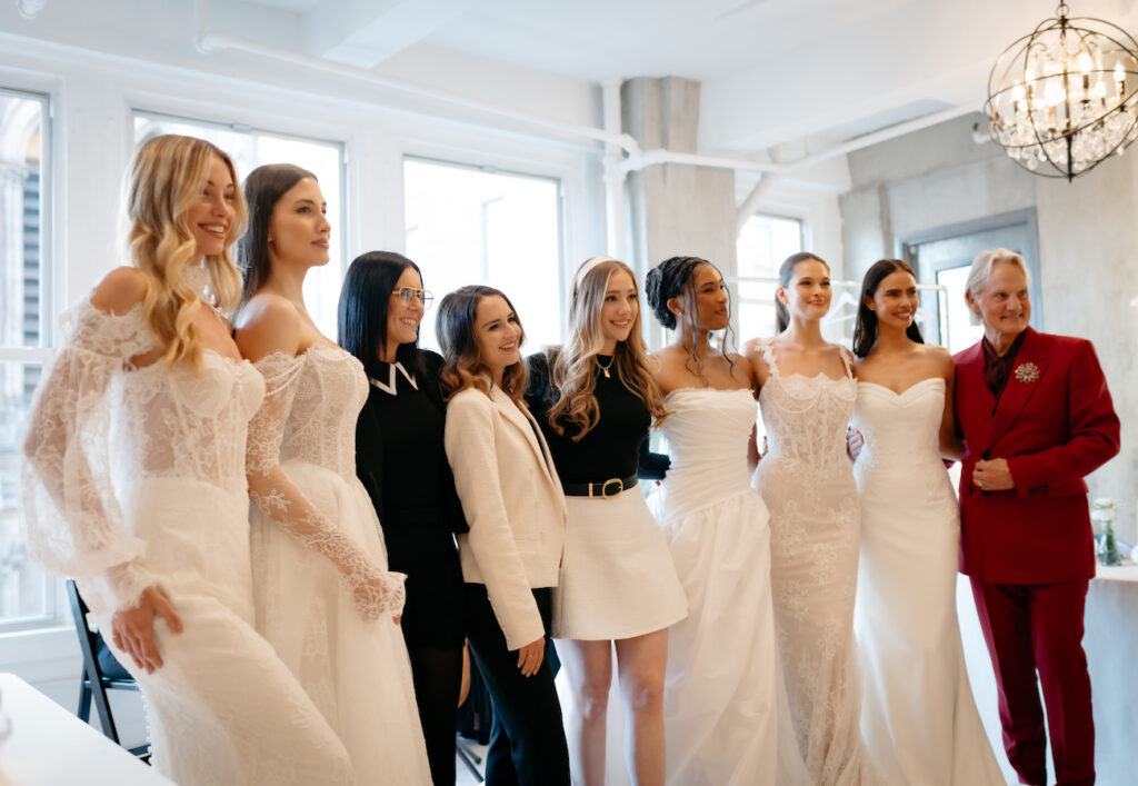 A group of women, some in wedding dresses and others in business attire, stand together and pose for a photo in a bright room.