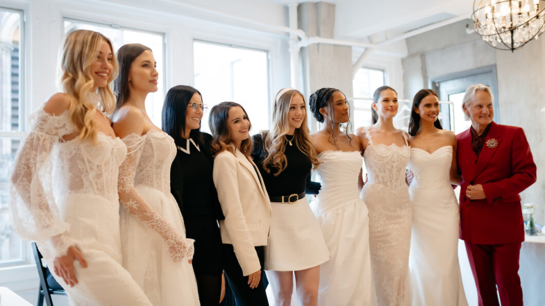 A group of women, some in wedding dresses and others in business attire, stand together and pose for a photo in a bright room.