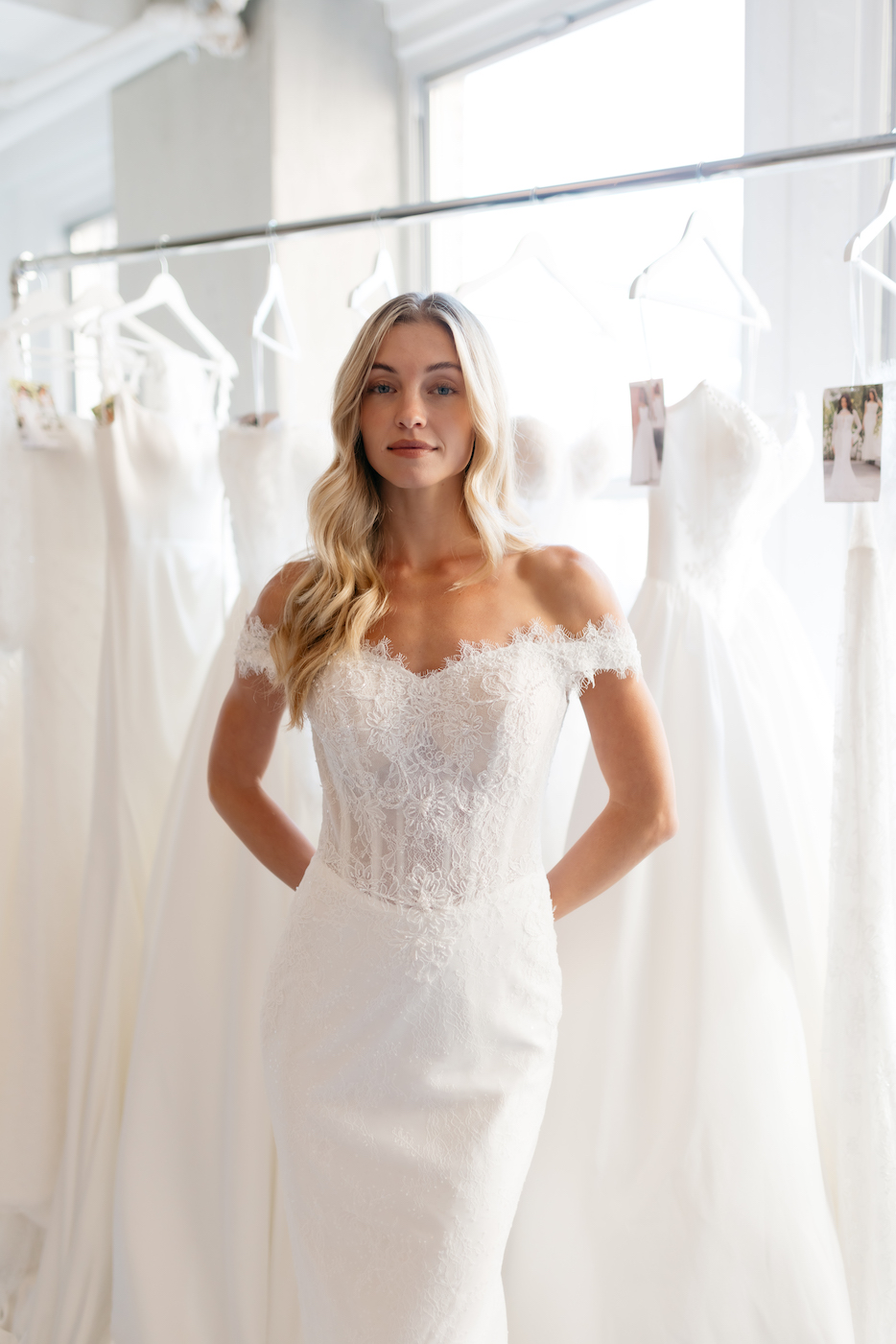 A woman wearing an off-the-shoulder lace wedding dress stands in front of a rack of white bridal gowns.