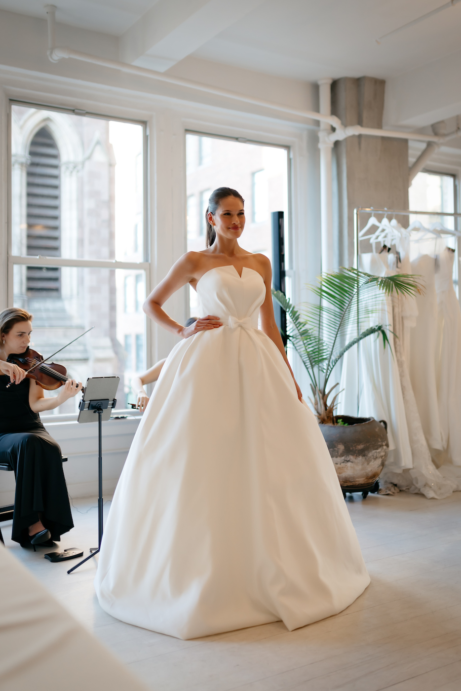 A woman wearing a strapless white ballgown-style wedding dress stands indoors, with a violinist playing in the background and wedding dresses hanging nearby.