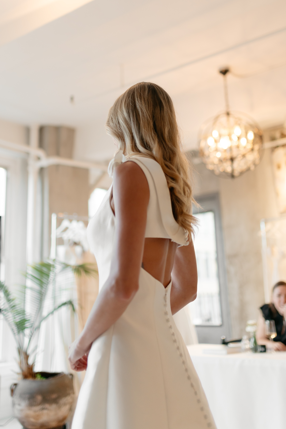 A woman in a white sleeveless wedding dress stands indoors near a window, with her back partially visible.