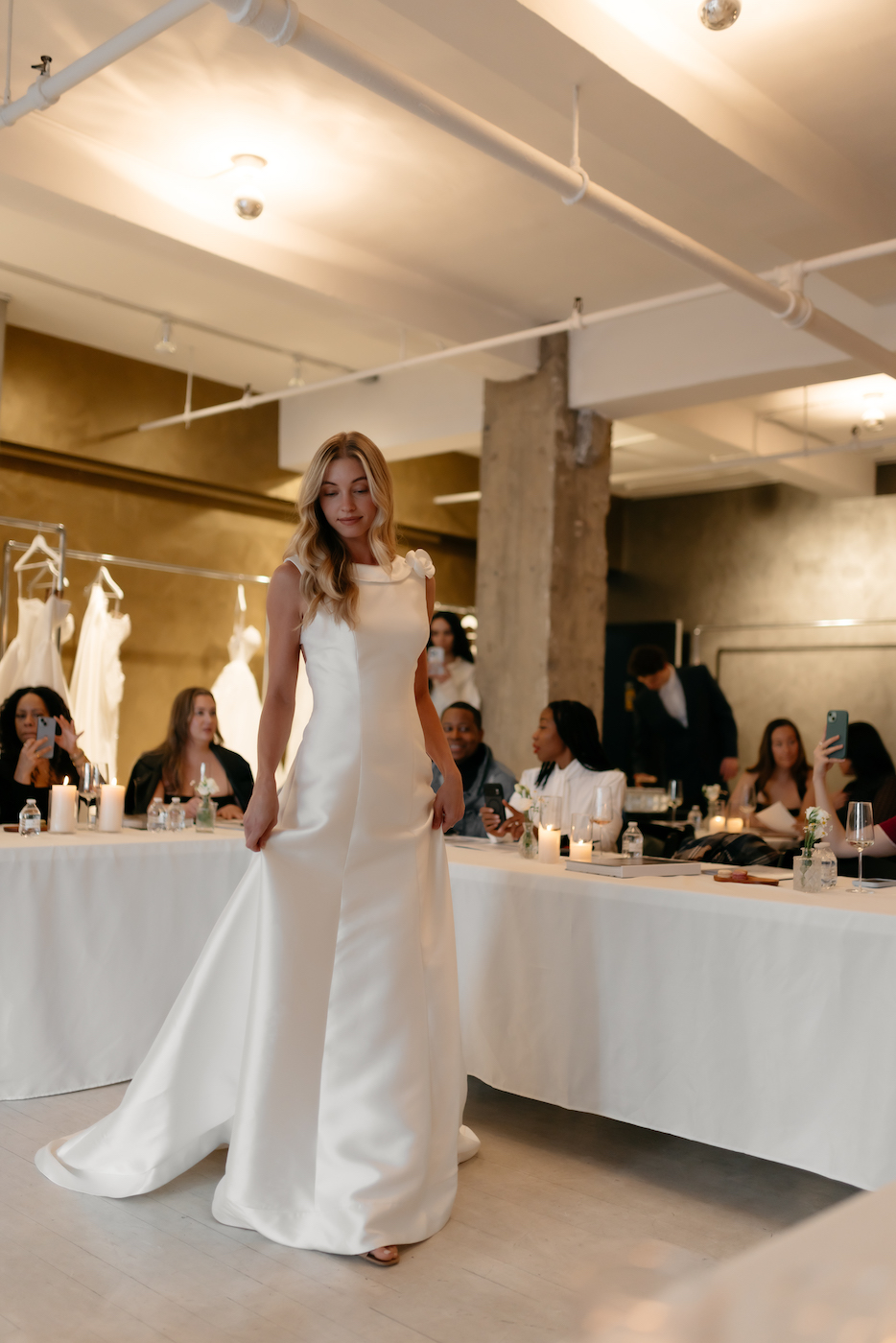 A woman in a white wedding dress stands in front of seated guests at an indoor event with wedding gowns hanging in the background.