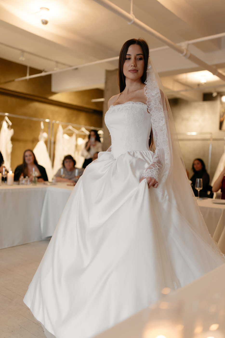A woman in a white wedding dress stands indoors, posing in front of seated people at what appears to be a bridal event or fitting.