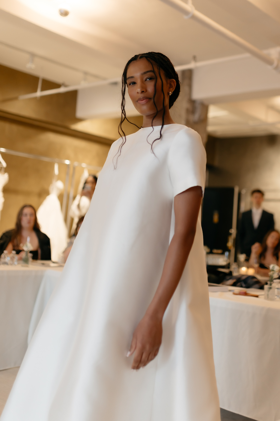 A woman wearing a white, short-sleeved, floor-length dress stands indoors at an event, with people seated at tables in the background.