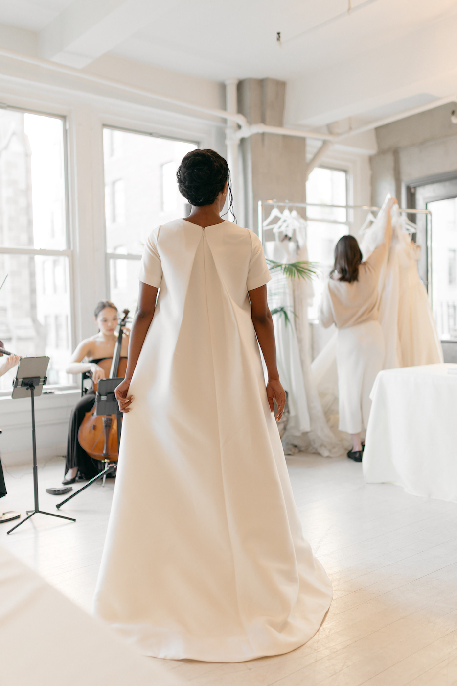 A person in a white gown stands indoors, facing away, while others adjust dresses and play musical instruments in the background.