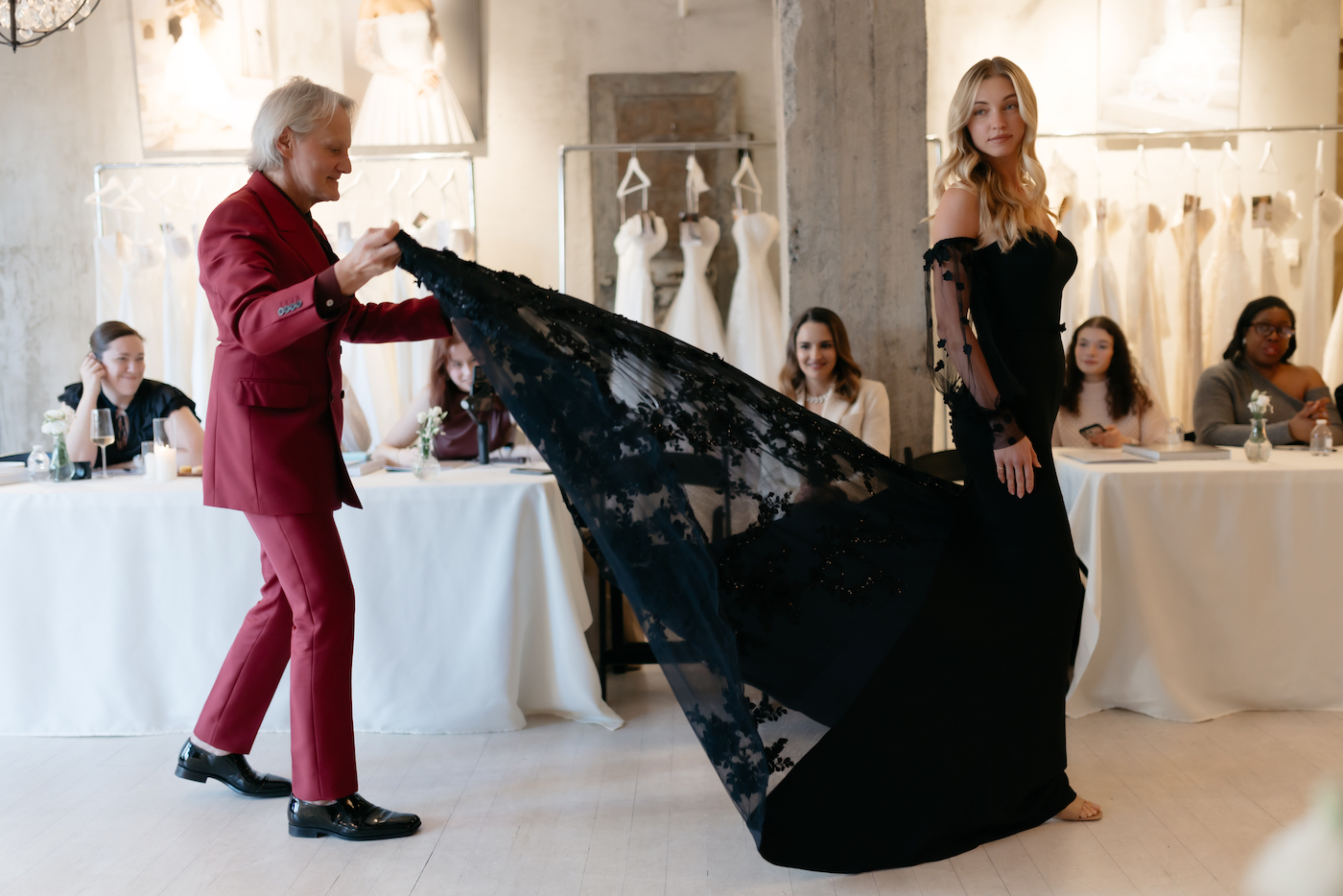 A person in a red suit holds the train of a woman's long black gown as she walks, with several seated people observing in a bridal boutique setting.