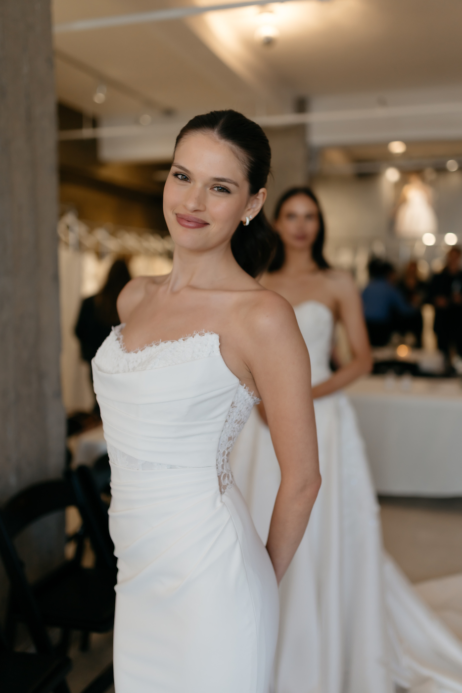 Two women in white strapless wedding dresses stand indoors, one in the foreground smiling at the camera, with another woman blurred in the background.