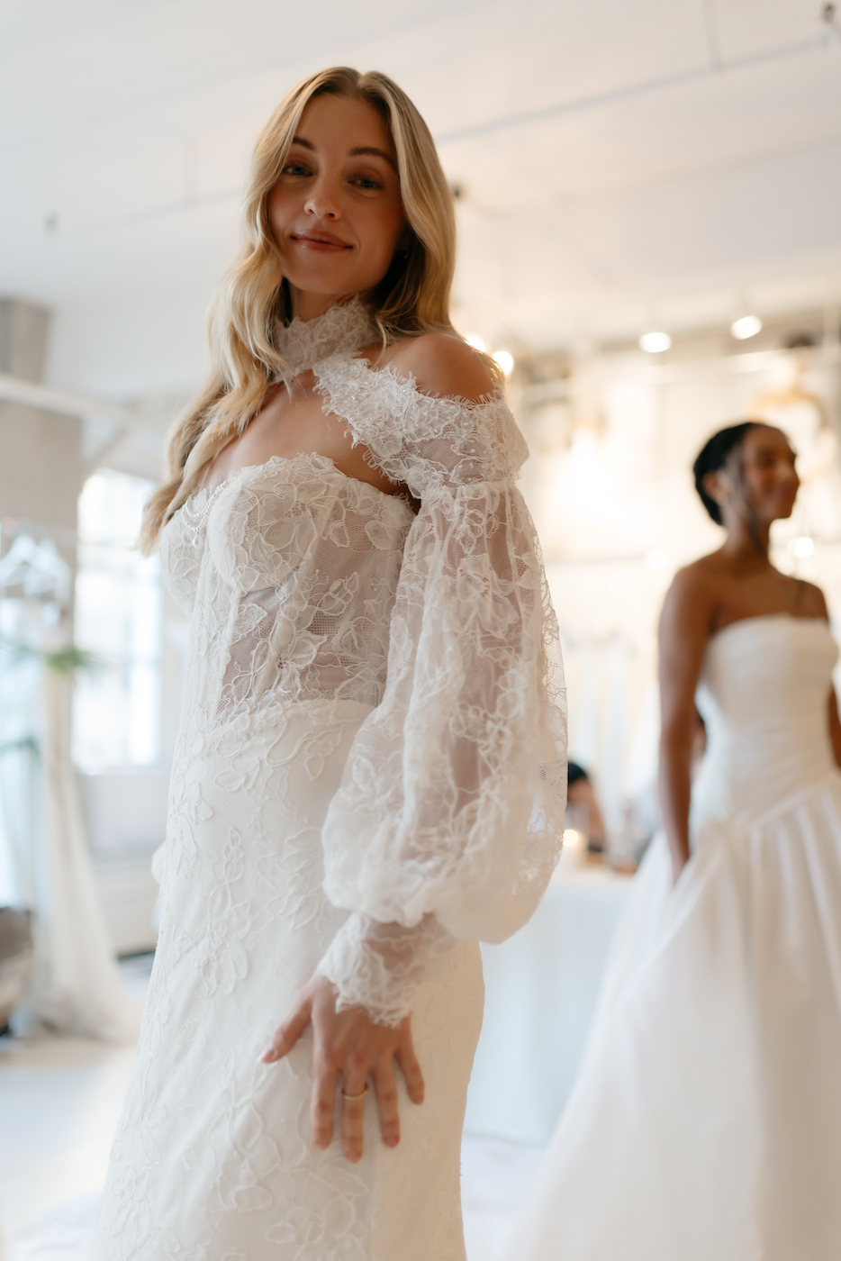 Two women wear white wedding dresses; the woman in the foreground has long, wavy hair and a lace dress with off-the-shoulder sleeves, while the other stands blurred in the background.