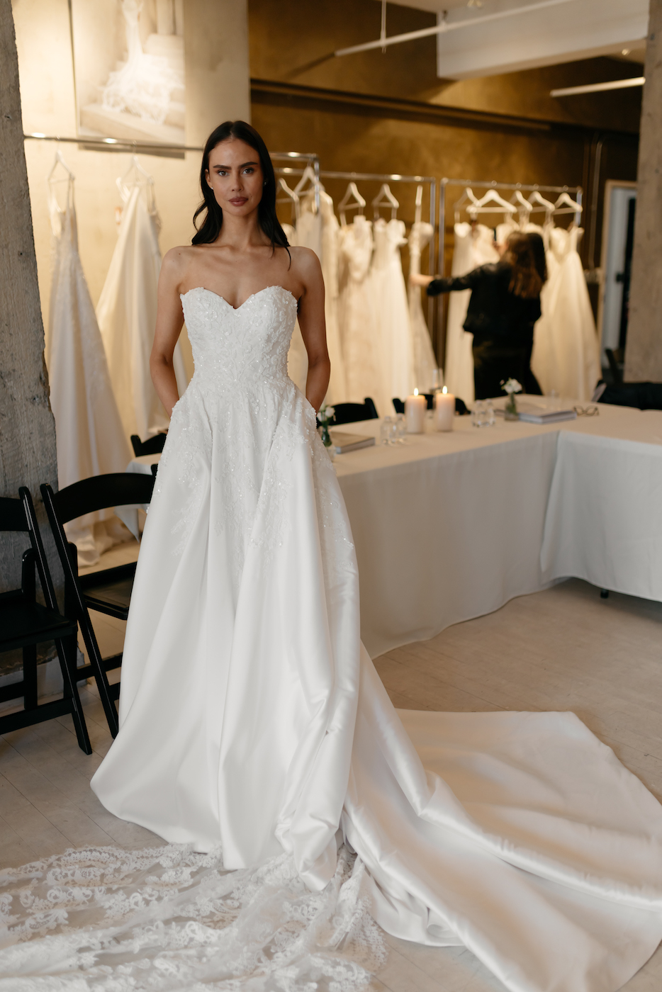 A woman wearing a strapless white wedding gown with a long train stands in a bridal shop, surrounded by hanging dresses.