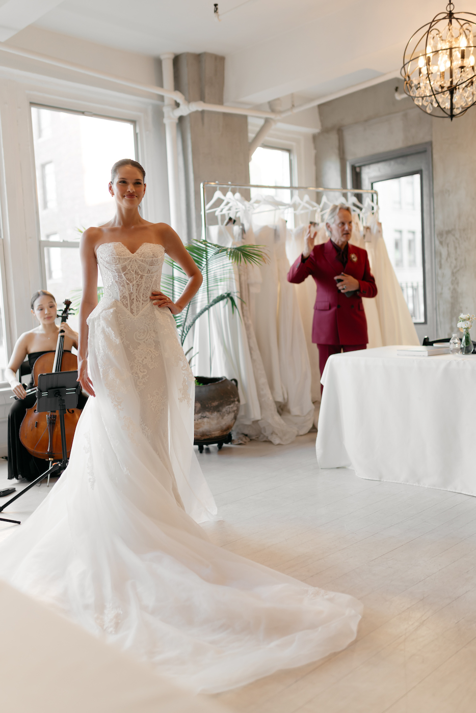 A bride in a strapless wedding gown stands in a bright room, with a cellist seated and a person in a red suit near racks of dresses in the background.