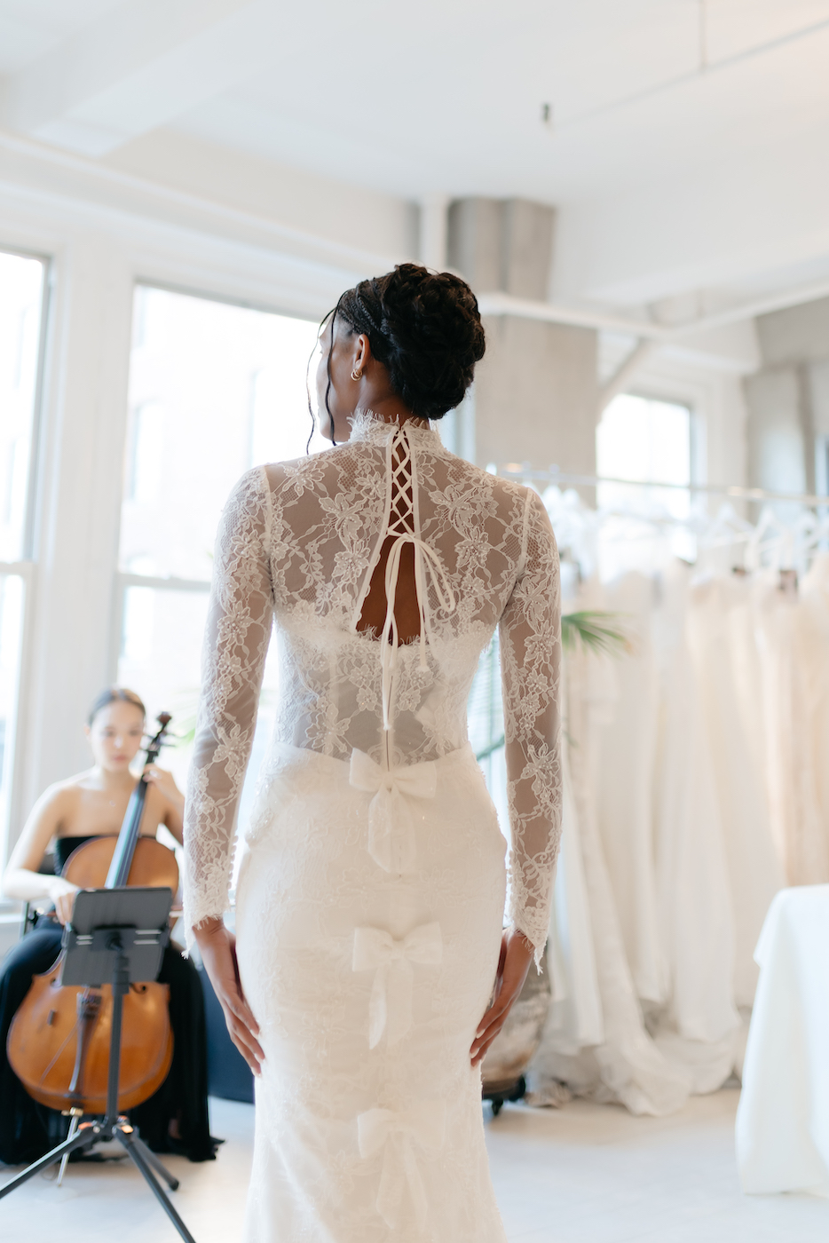 A woman in a lace-back white wedding dress stands indoors, facing away; a seated cellist and a rack of dresses are visible in the background.