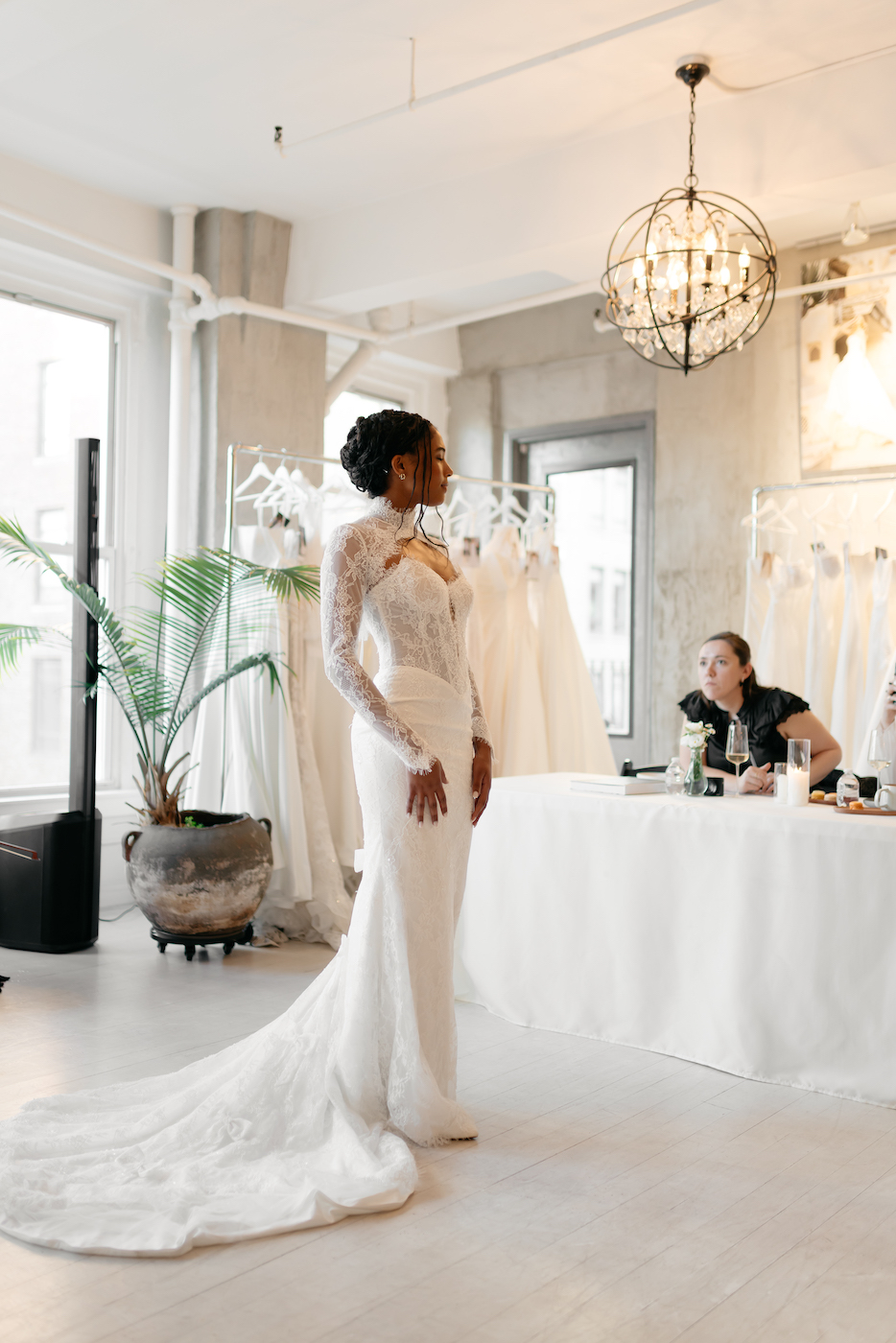 A woman in a long-sleeved wedding dress stands in a bridal shop, while two people seated at a table observe her.