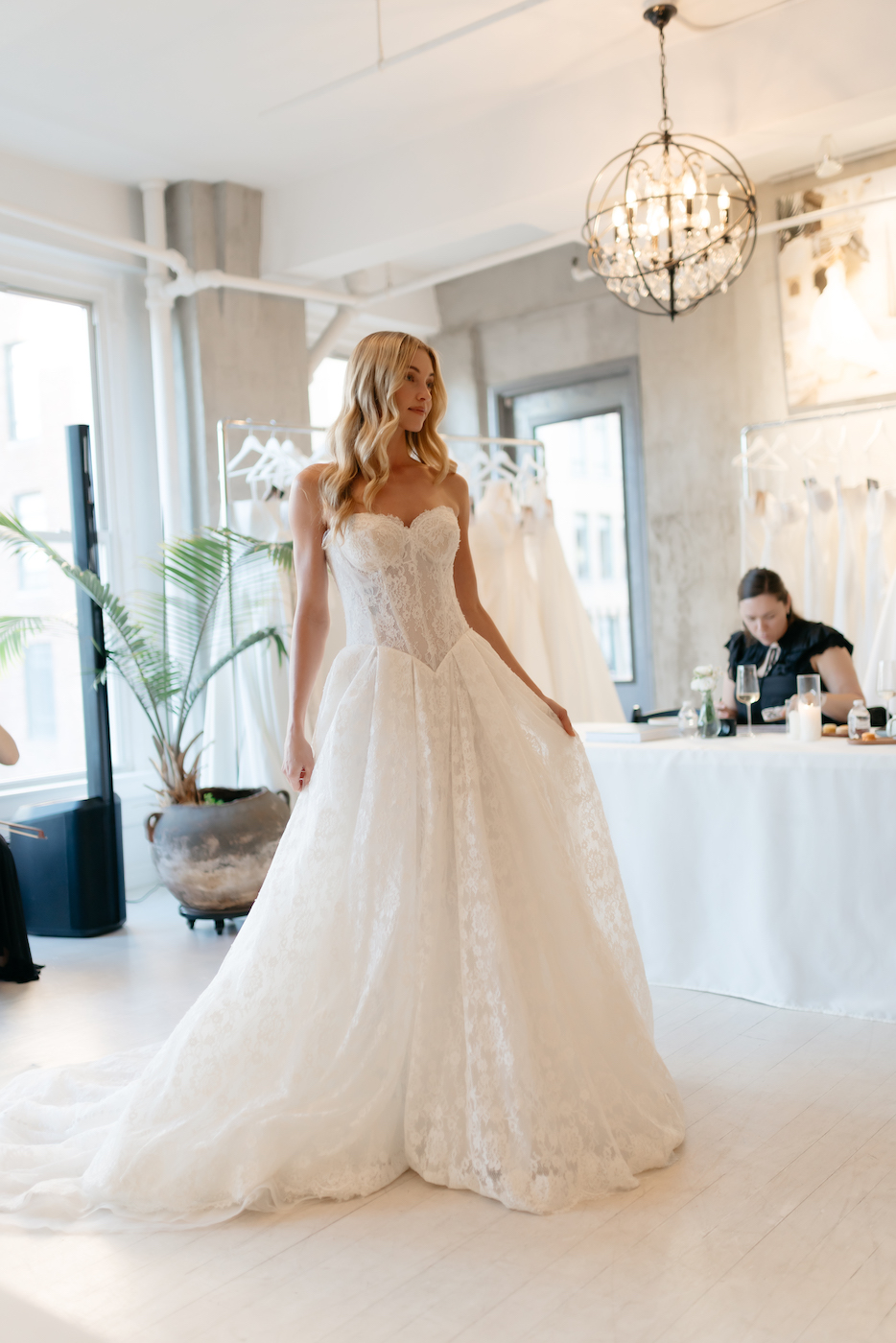 A woman wearing a strapless white wedding dress stands in a bright bridal boutique as another person sits at a table in the background.
