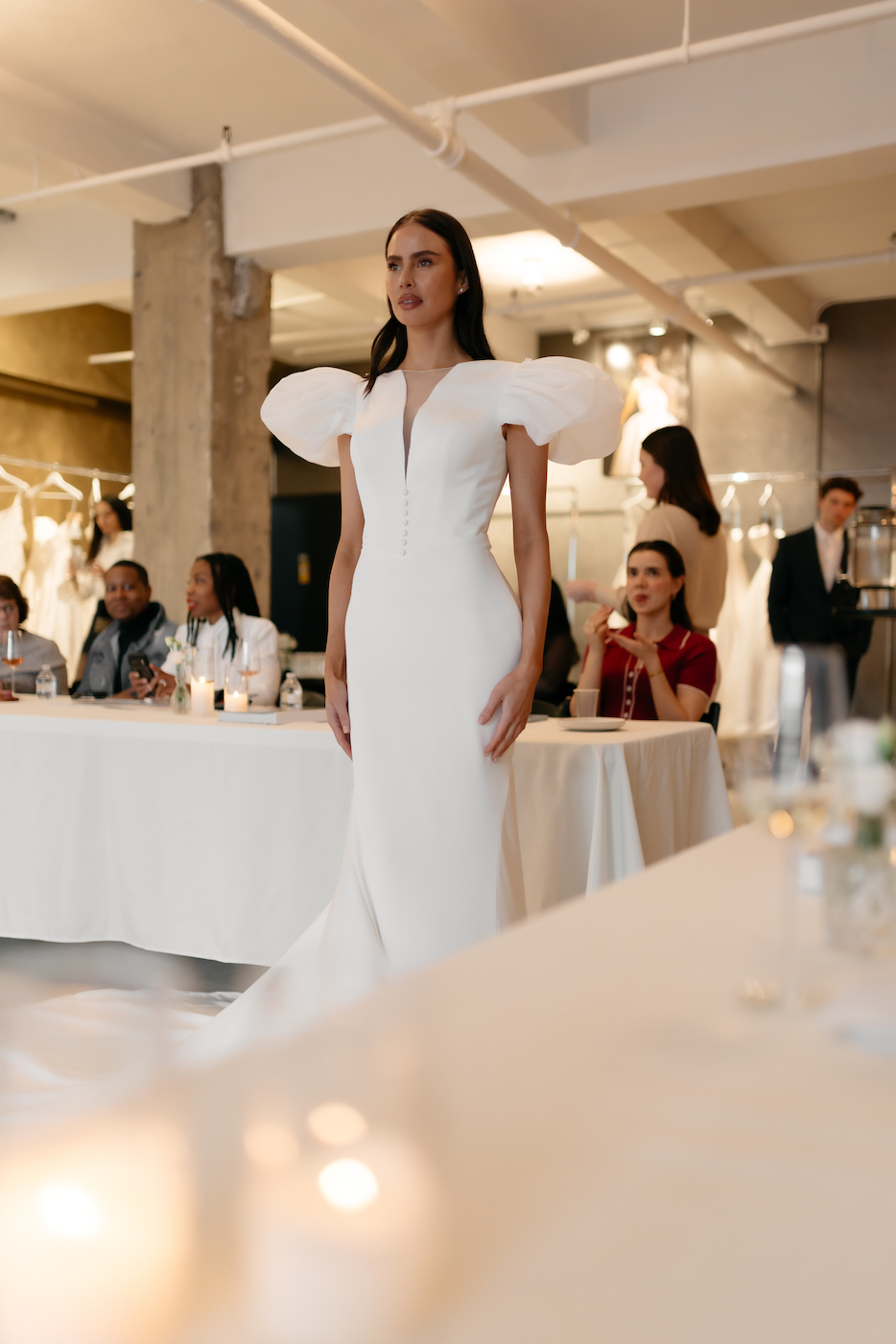 A woman in a white gown with dramatic puffed sleeves walks indoors while seated people watch from tables in a well-lit room.