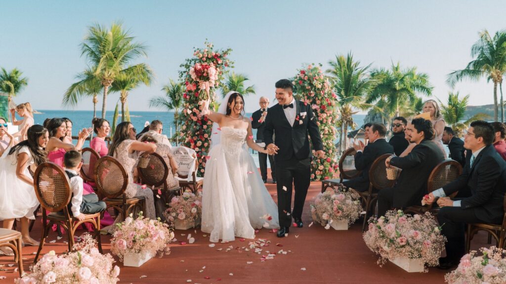 A bride and groom walk down an outdoor aisle lined with flowers, smiling and holding hands, surrounded by guests at a tropical wedding ceremony.