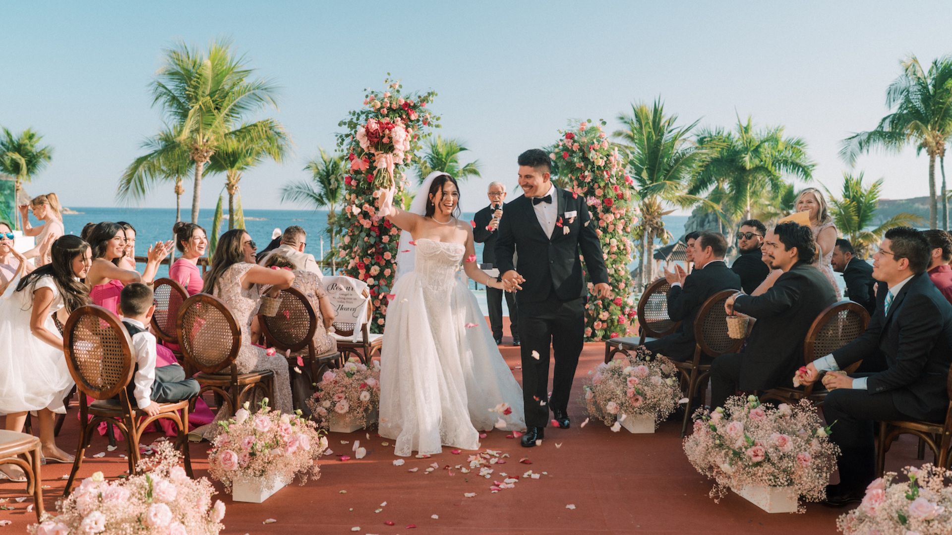A bride and groom walk down an outdoor aisle lined with flowers, smiling and holding hands, surrounded by guests at a tropical wedding ceremony.