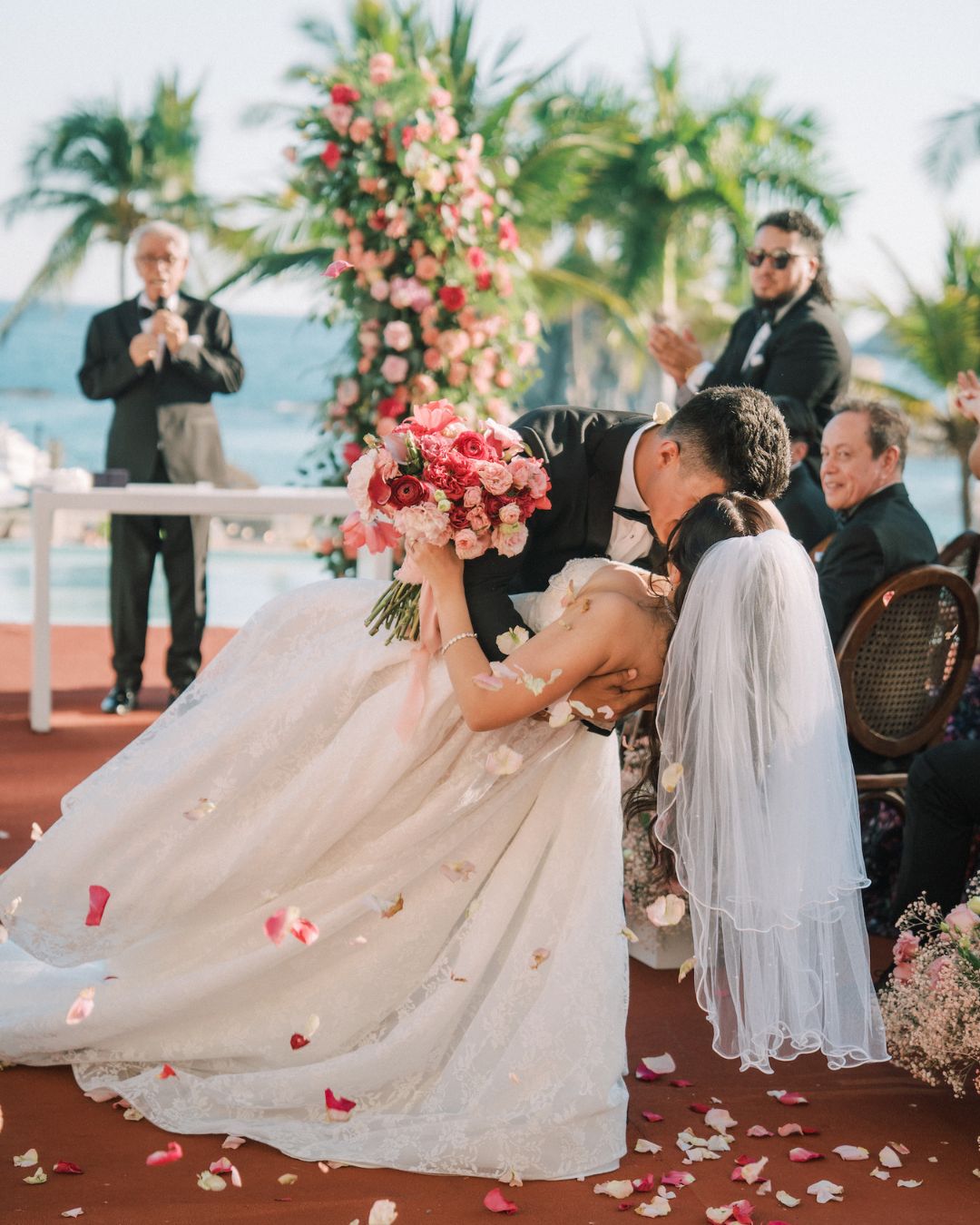 A bride and groom kiss during their wedding ceremony outdoors, surrounded by guests, flower petals, palm trees, and a floral arch.