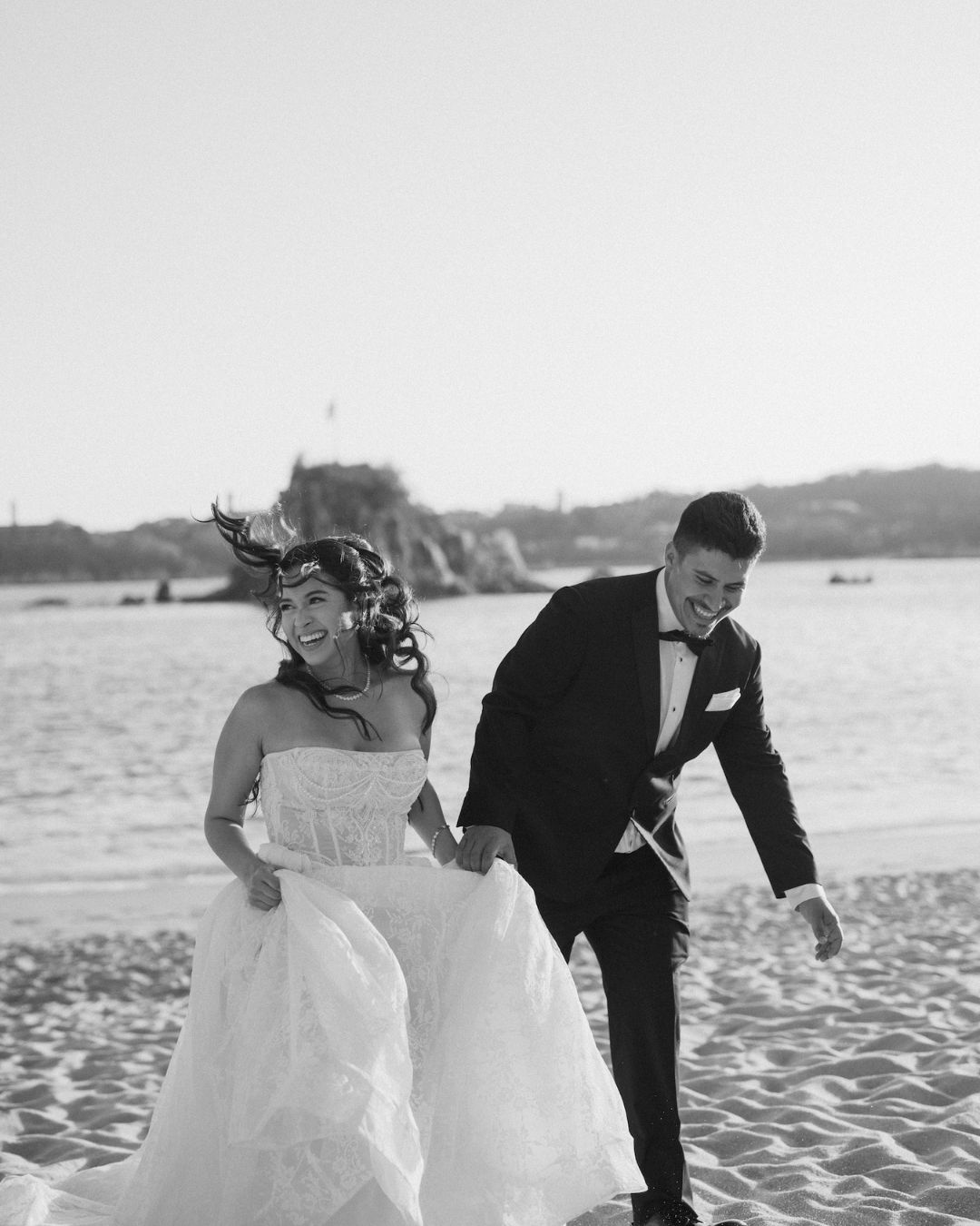 A bride and groom in formal wedding attire walk hand in hand on a sandy beach, smiling and laughing.