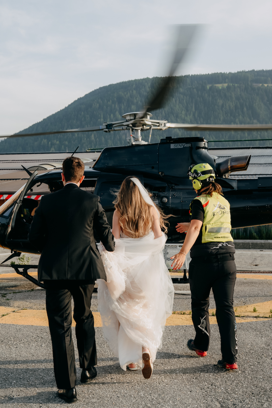 A bride and groom walk toward a helicopter, accompanied by a crew member in safety gear, with mountains in the background.