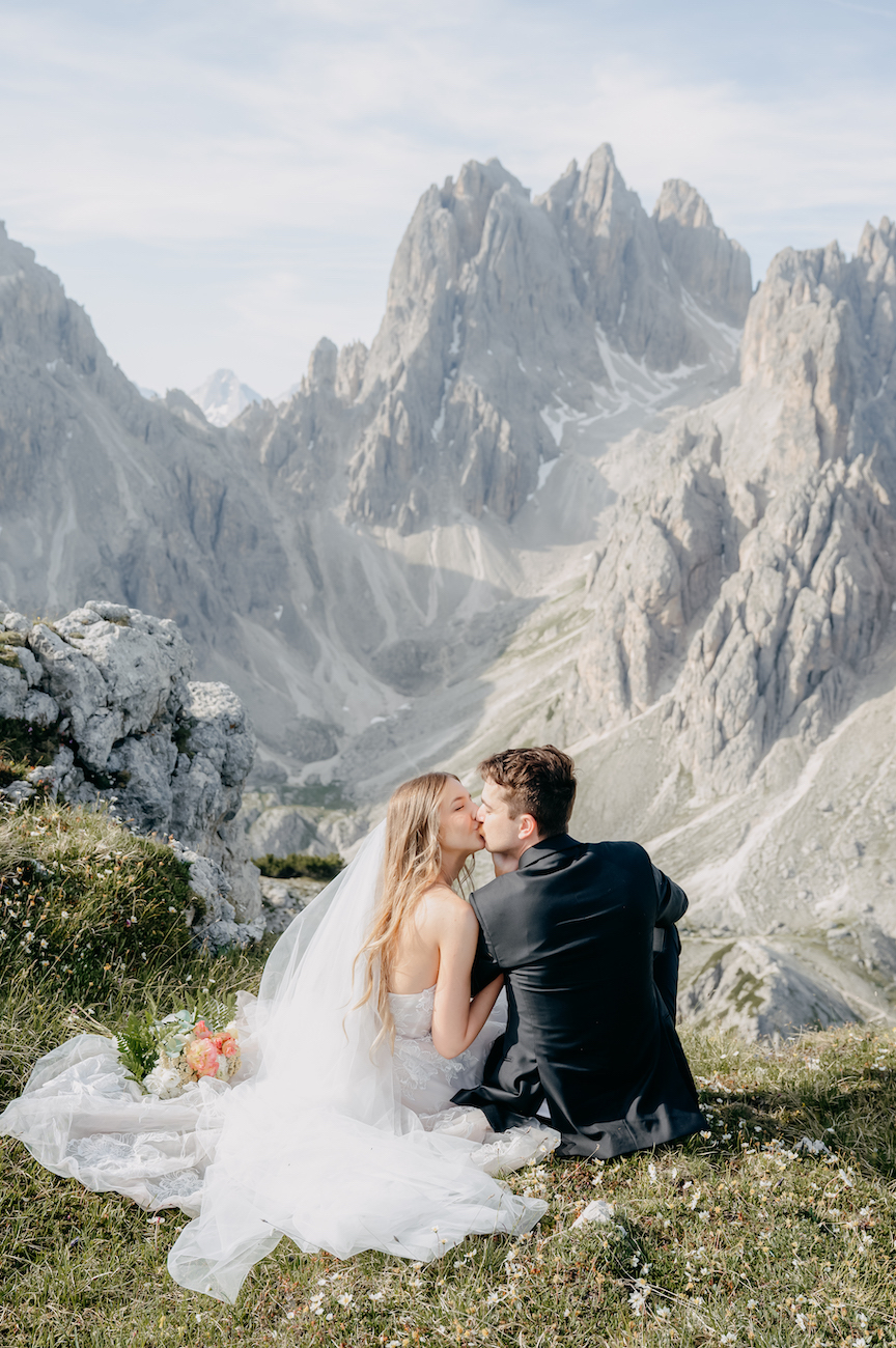 A bride and groom sit on grass, kissing, with rocky mountain peaks in the background.