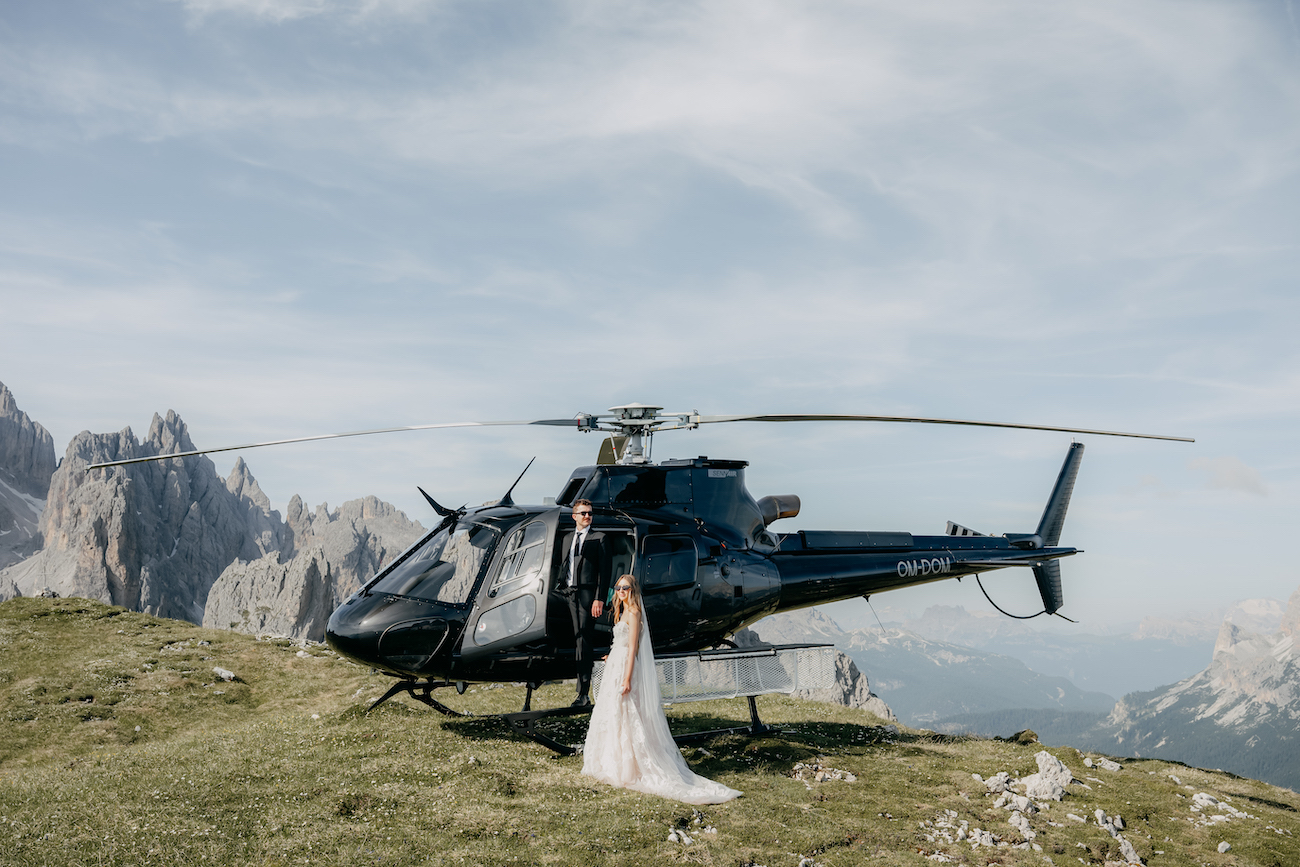 A bride and groom stand beside a black helicopter on a grassy mountain plateau with rocky peaks in the background.