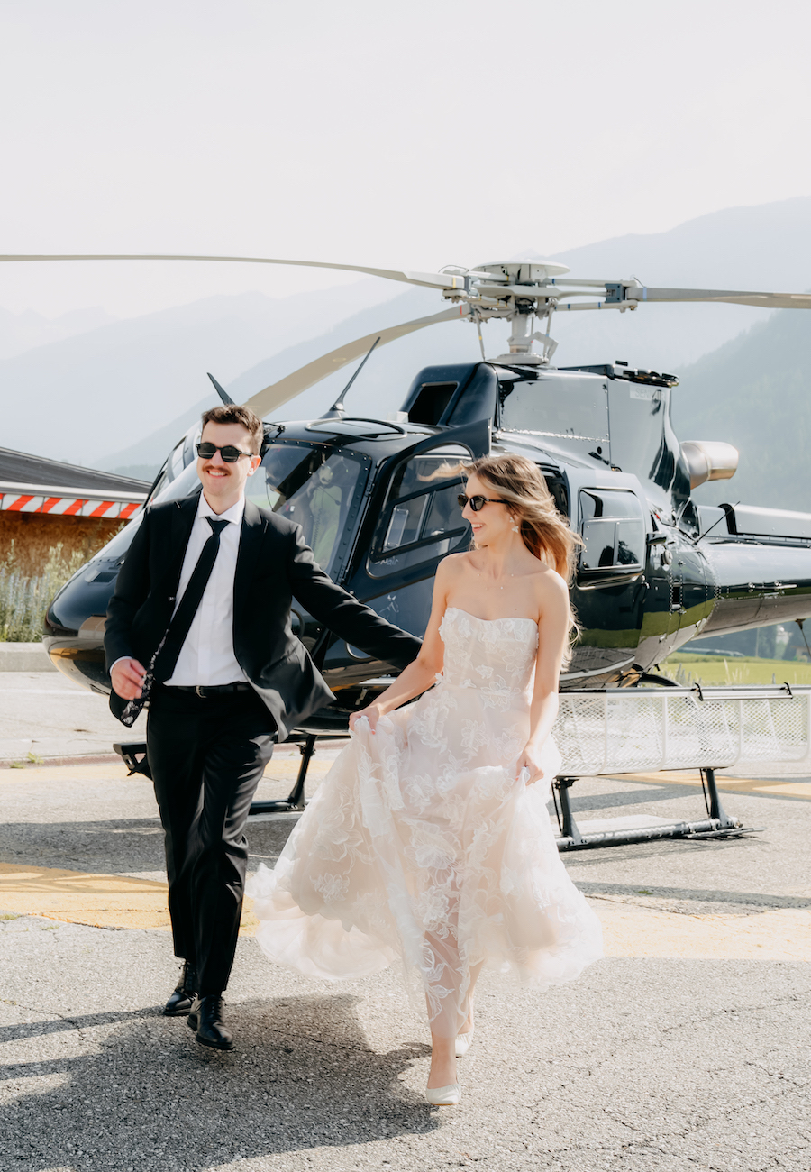A man in a suit and a woman in a wedding dress walk hand in hand on a helipad in front of a helicopter.
