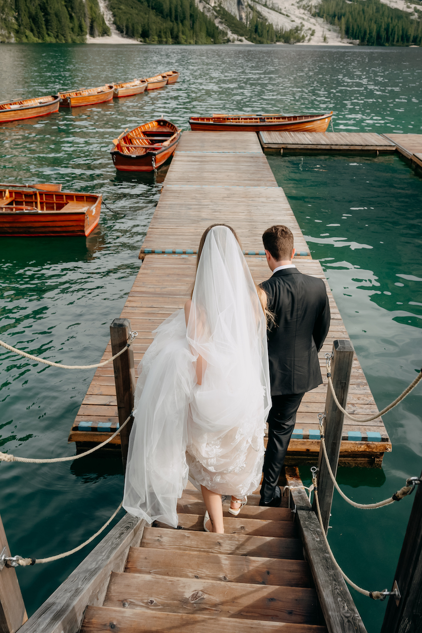 A bride and groom walk down wooden steps toward a dock with boats on a lake, surrounded by trees and mountains.
