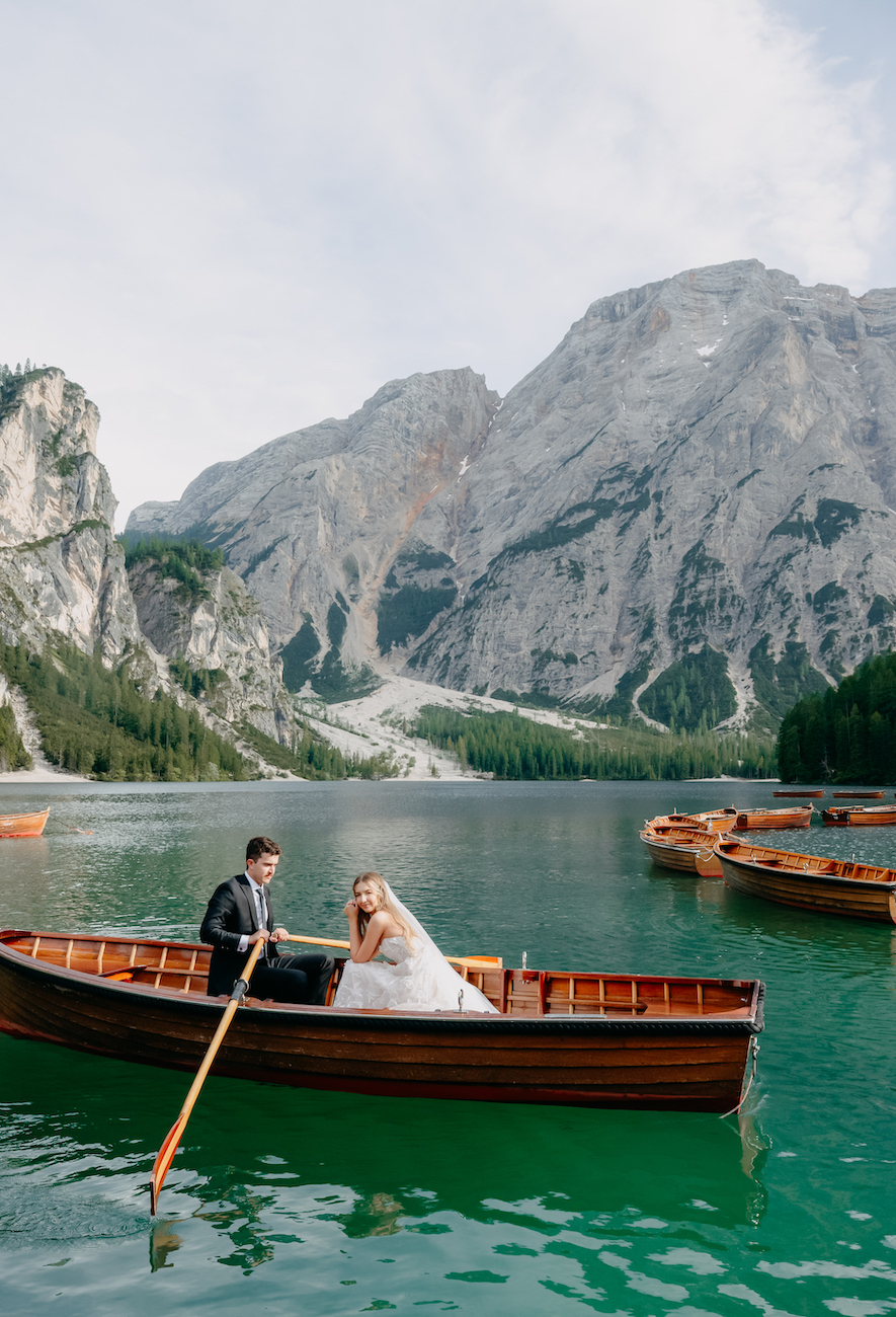 A bride and groom sit in a wooden rowboat on a mountain lake, surrounded by other boats, with rocky mountains and pine trees in the background.