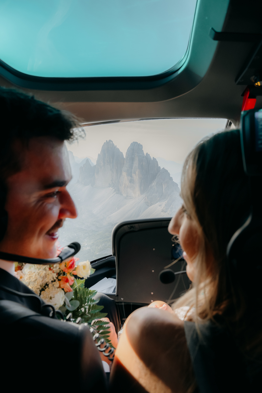 A man and a woman, both wearing headsets, sit in a helicopter with mountains visible through the window in the background.