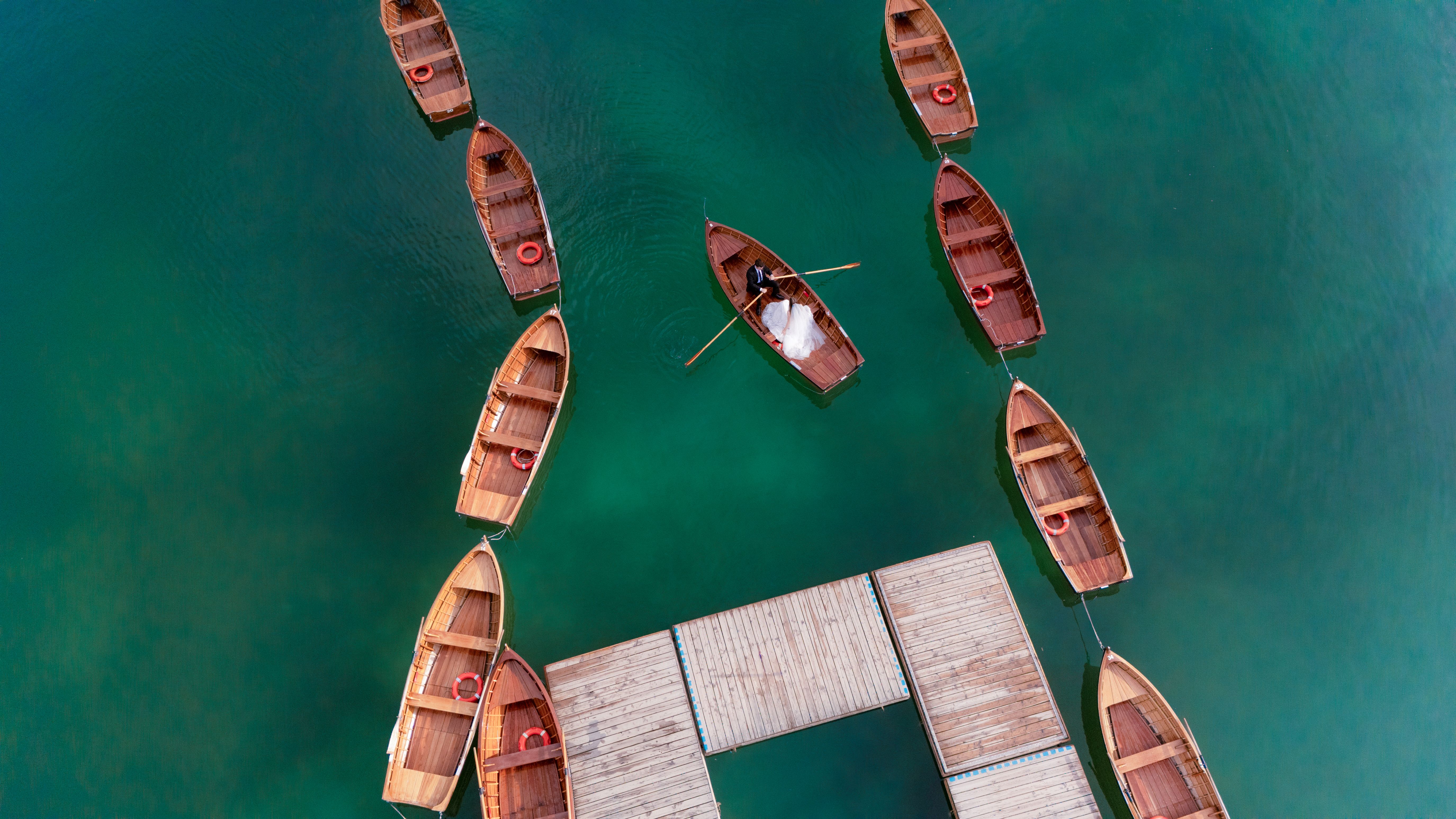 Aerial view of several wooden rowboats surrounding a dock on clear green water, with one boat being rowed near the center.