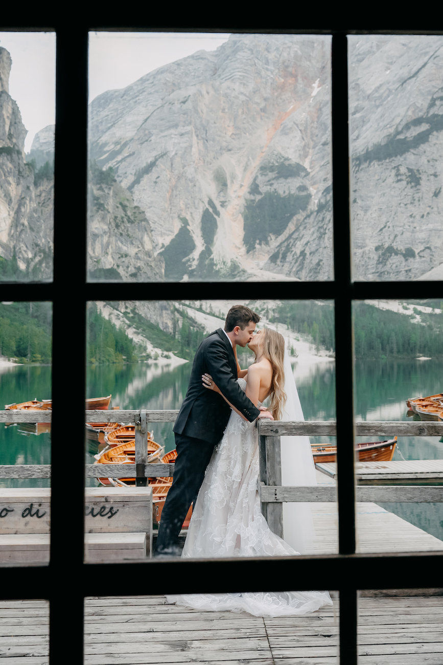 A bride and groom embrace and kiss on a wooden dock by a lake, with mountains and boats in the background, seen through a window.